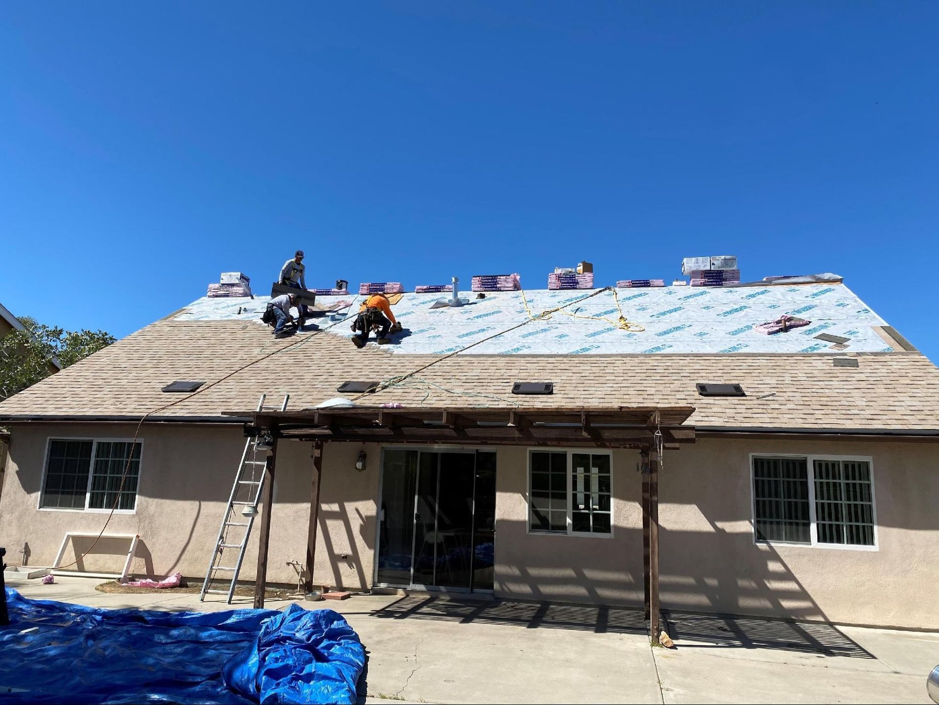 Workers install protective underlayment on a residential roof under a bright blue sky.
