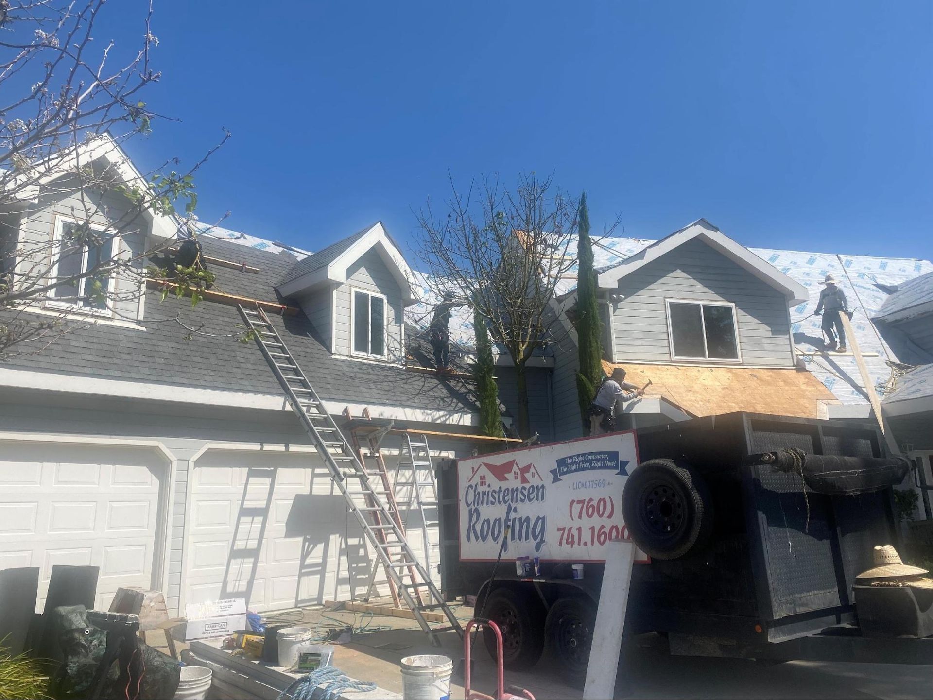 Roofers work on a house exterior with a dumpster in the driveway and a ladder against the roof.