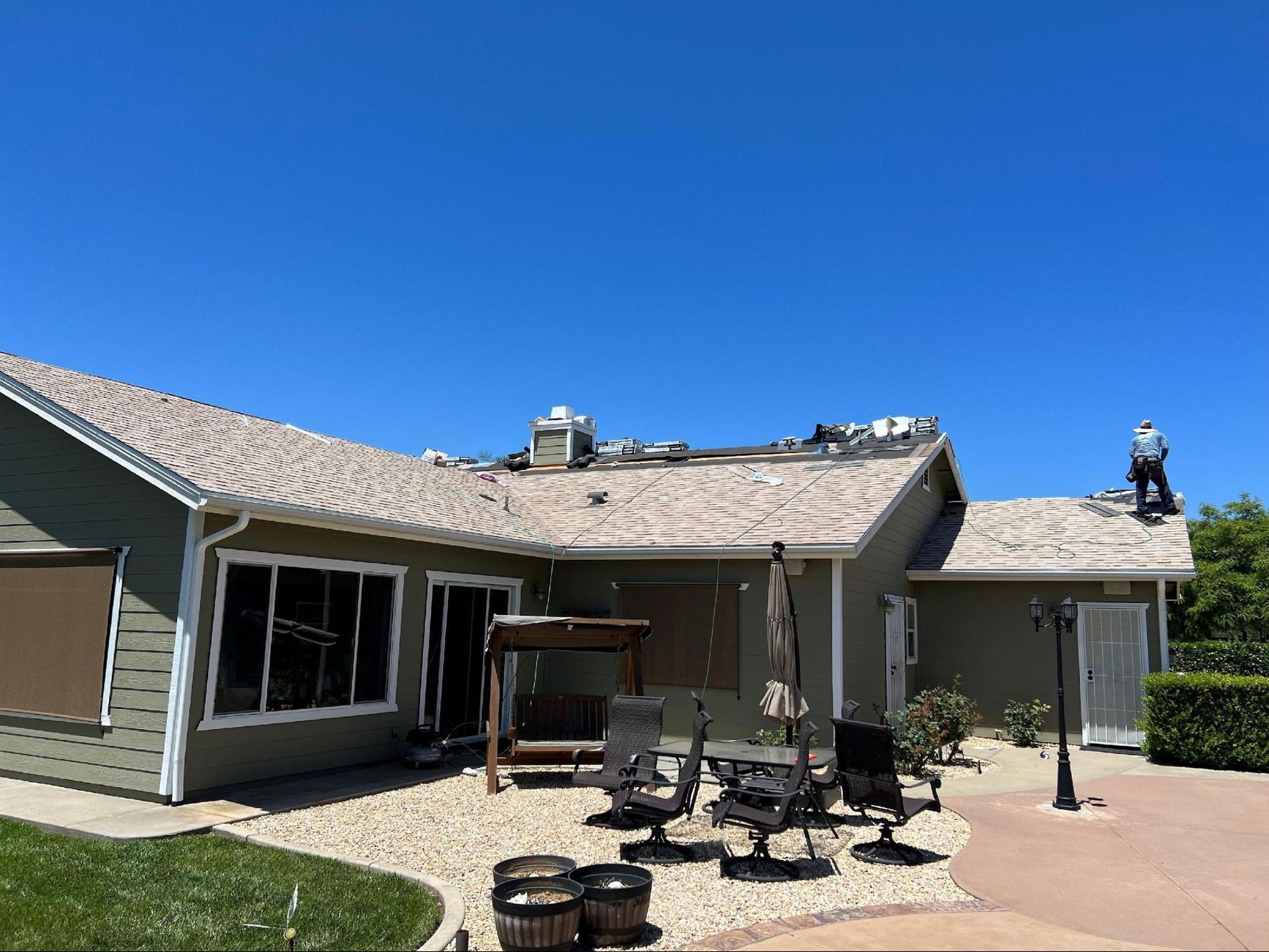 A worker performs roof repairs on a one-story olive green house on a sunny day with a patio area in the foreground.