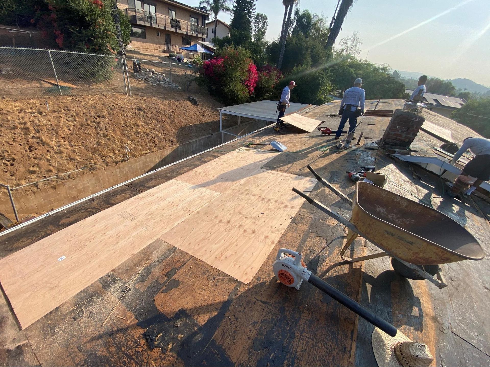 Workers replace plywood sheathing on a sunny roof, with a wheelbarrow and leaf blower nearby.