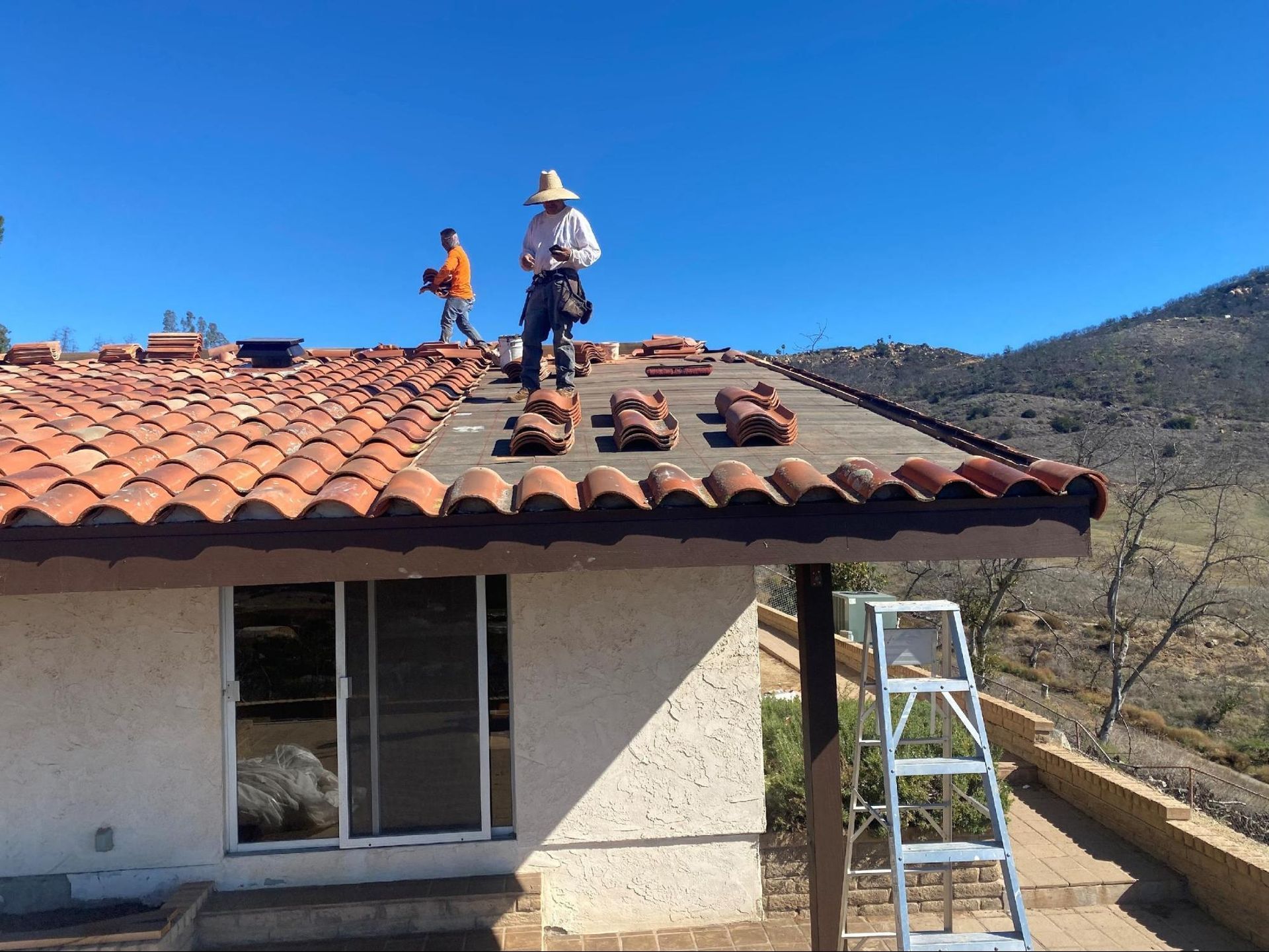 Two workers repair a tiled roof on a residential home in a desert landscape under a clear blue sky.