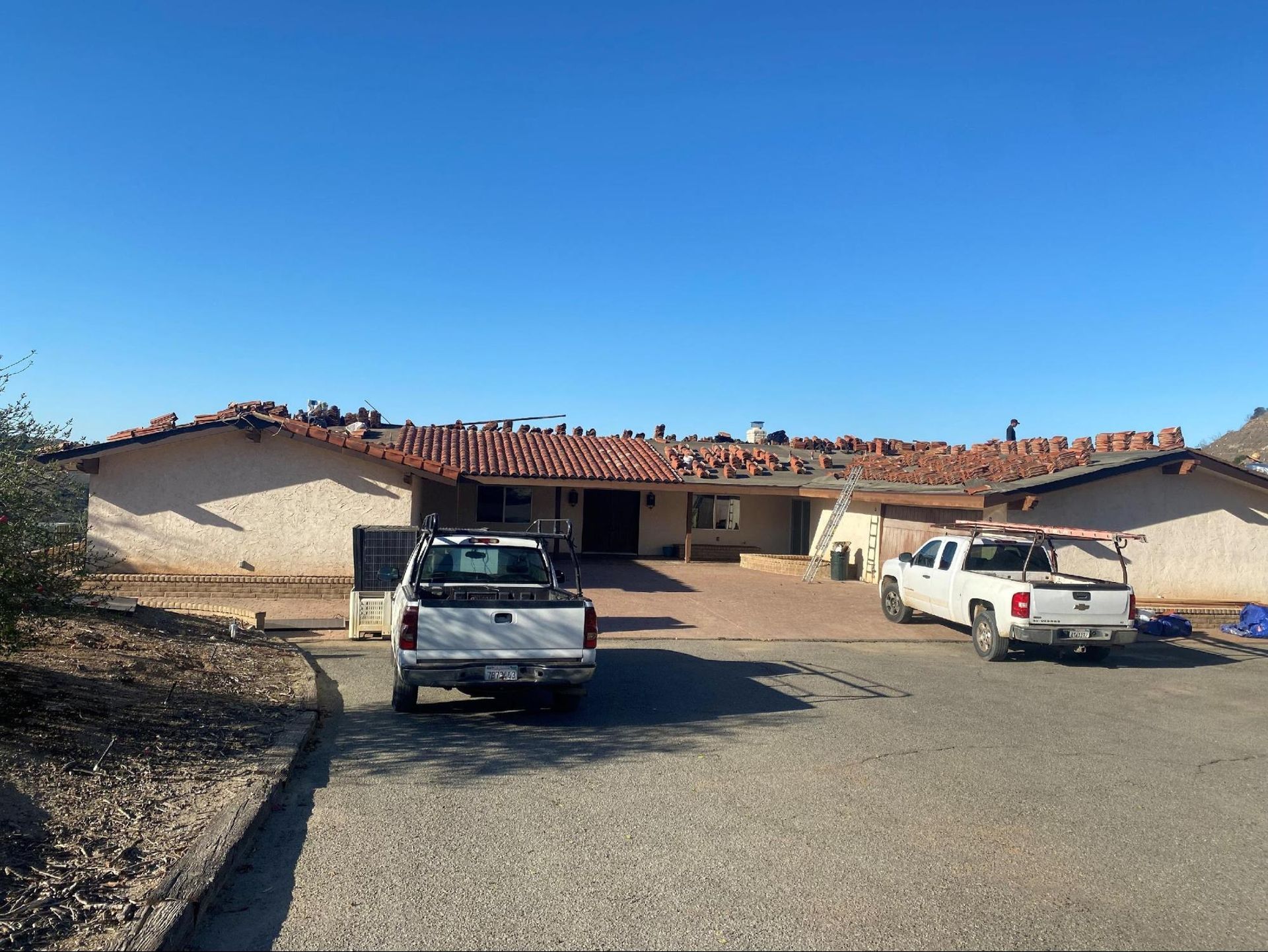 A single-story ranch-style home with a red-tiled roof and a gravel driveway featuring two white pickup trucks.