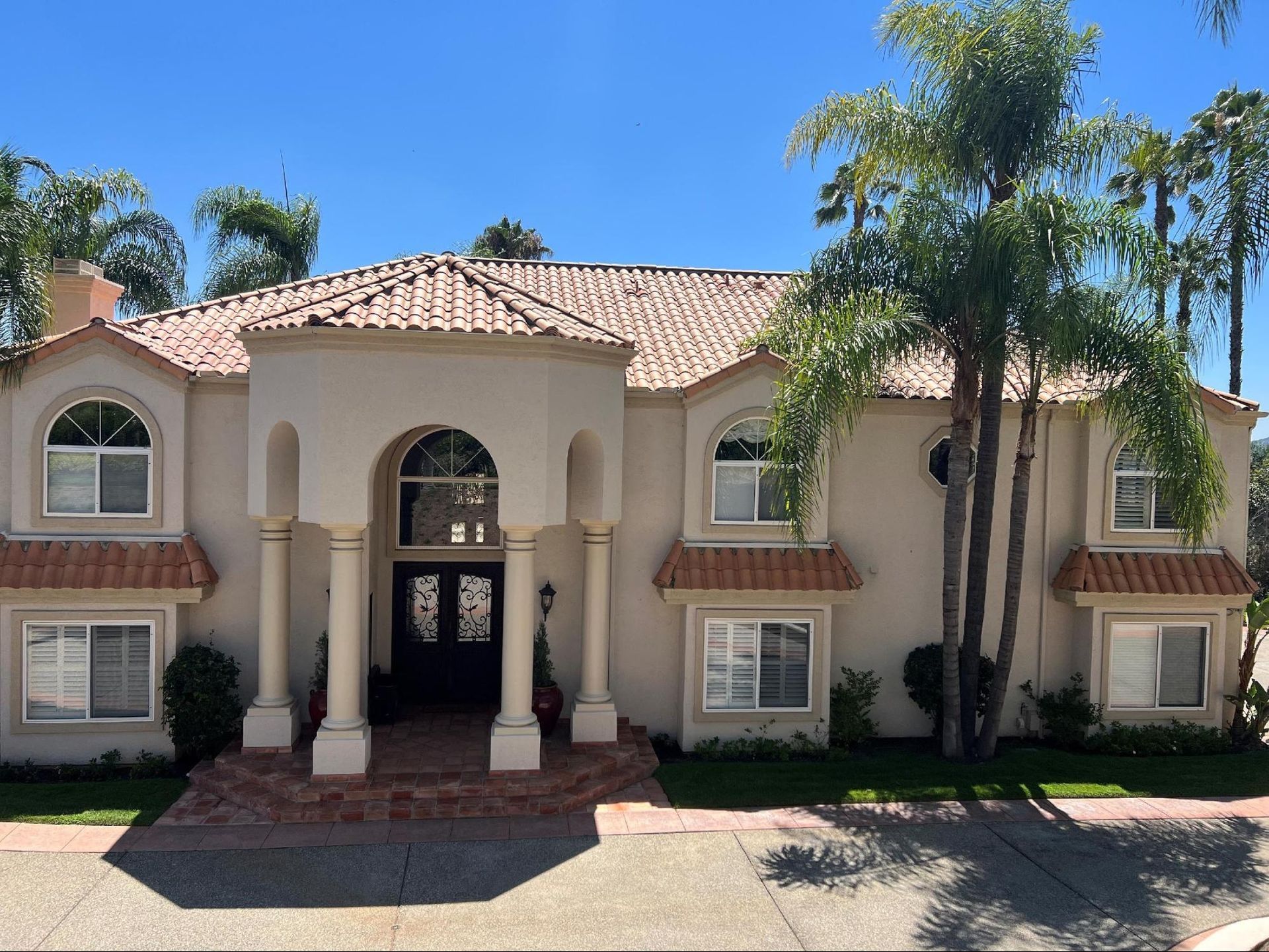 A two-story, beige stucco house with a tiled roof, arched entryway with pillars, and tall palm trees under a blue sky.