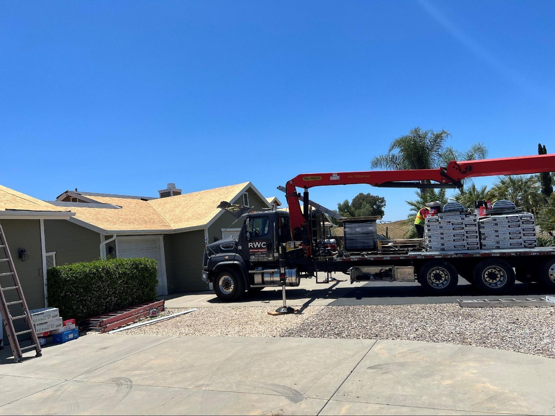 A crane truck parked on a residential driveway delivering pallets of roofing shingles to a house with a new roof underlay.