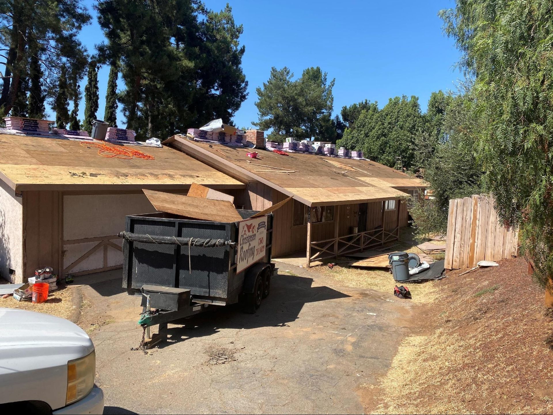 A house undergoing a roof replacement with exposed wooden decking, a trailer in the driveway, and trees in the background.