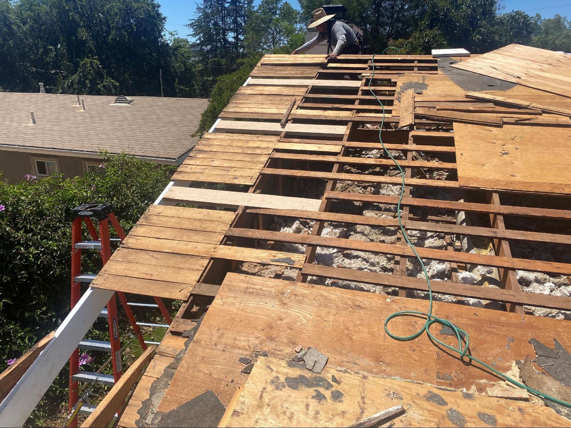 A construction worker stands on a partially torn-off roof, removing wooden slats to expose the rafters.