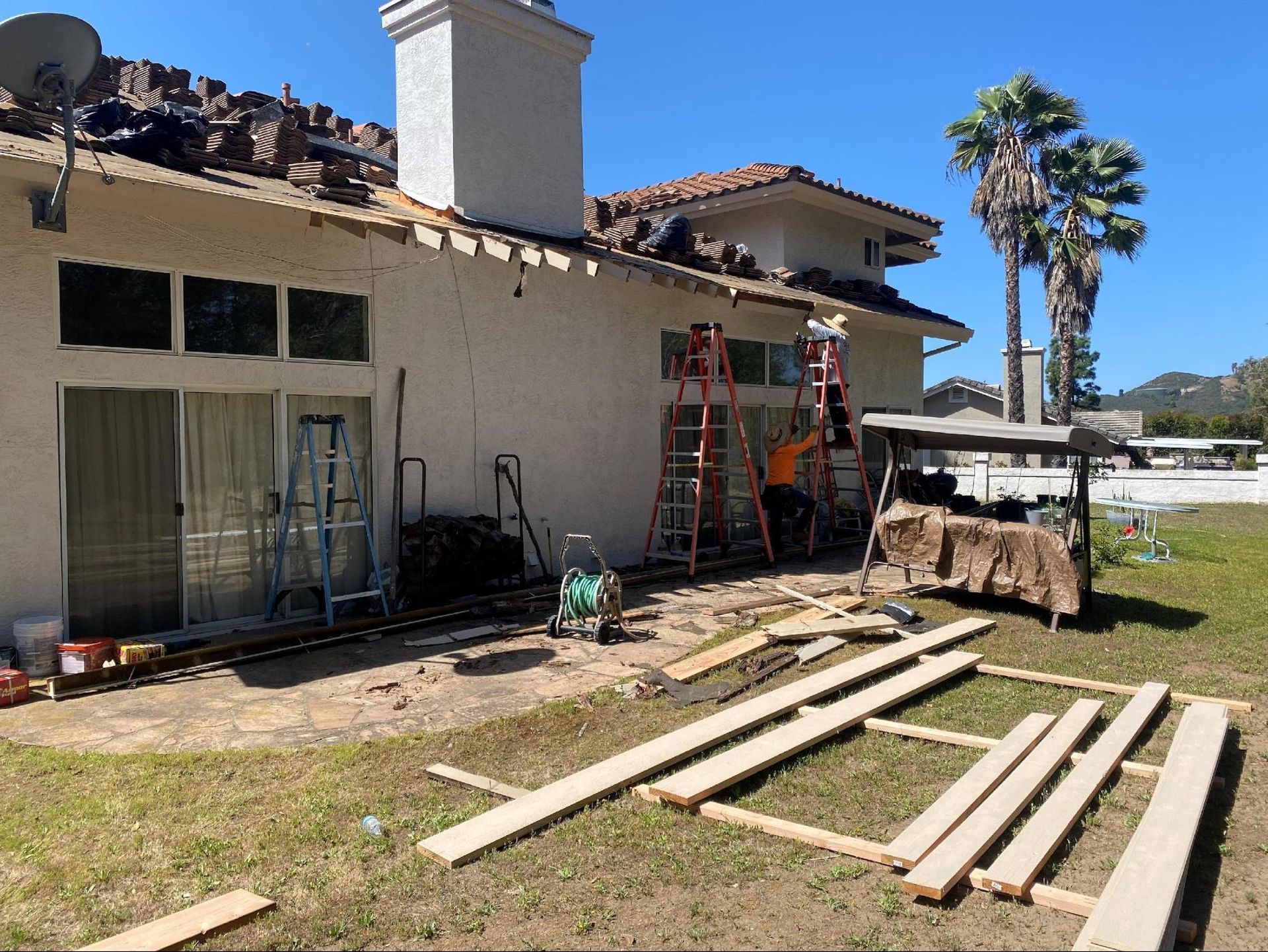 Construction workers on ladders perform roof repairs on a stucco house exterior with wooden boards laid on the lawn.