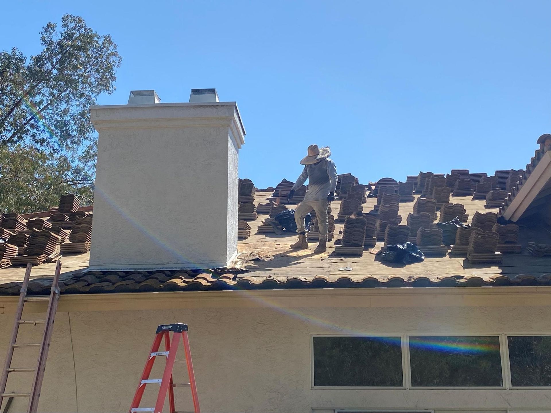 A worker wearing a protective suit stands on a residential roof removing clay tiles near a large white chimney.
