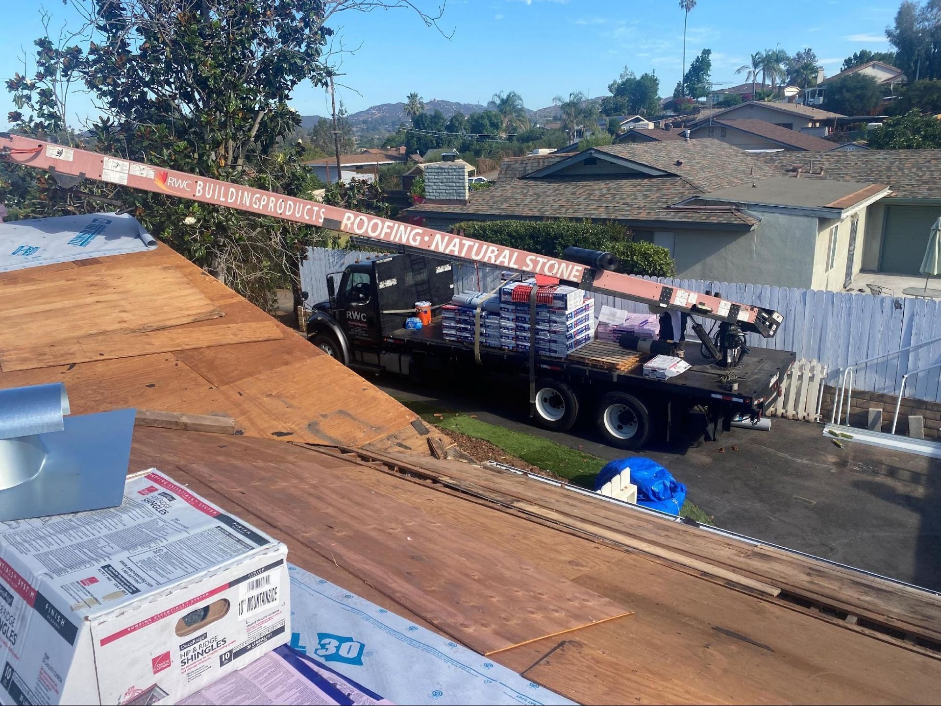 A flatbed delivery truck with a crane parked by a house under roof renovation, showing materials on a wooden roof deck.