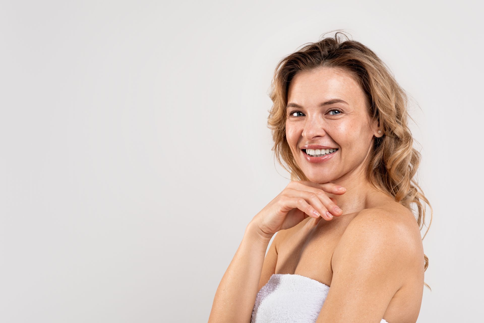 A woman wrapped in a white towel is smiling and touching her face.