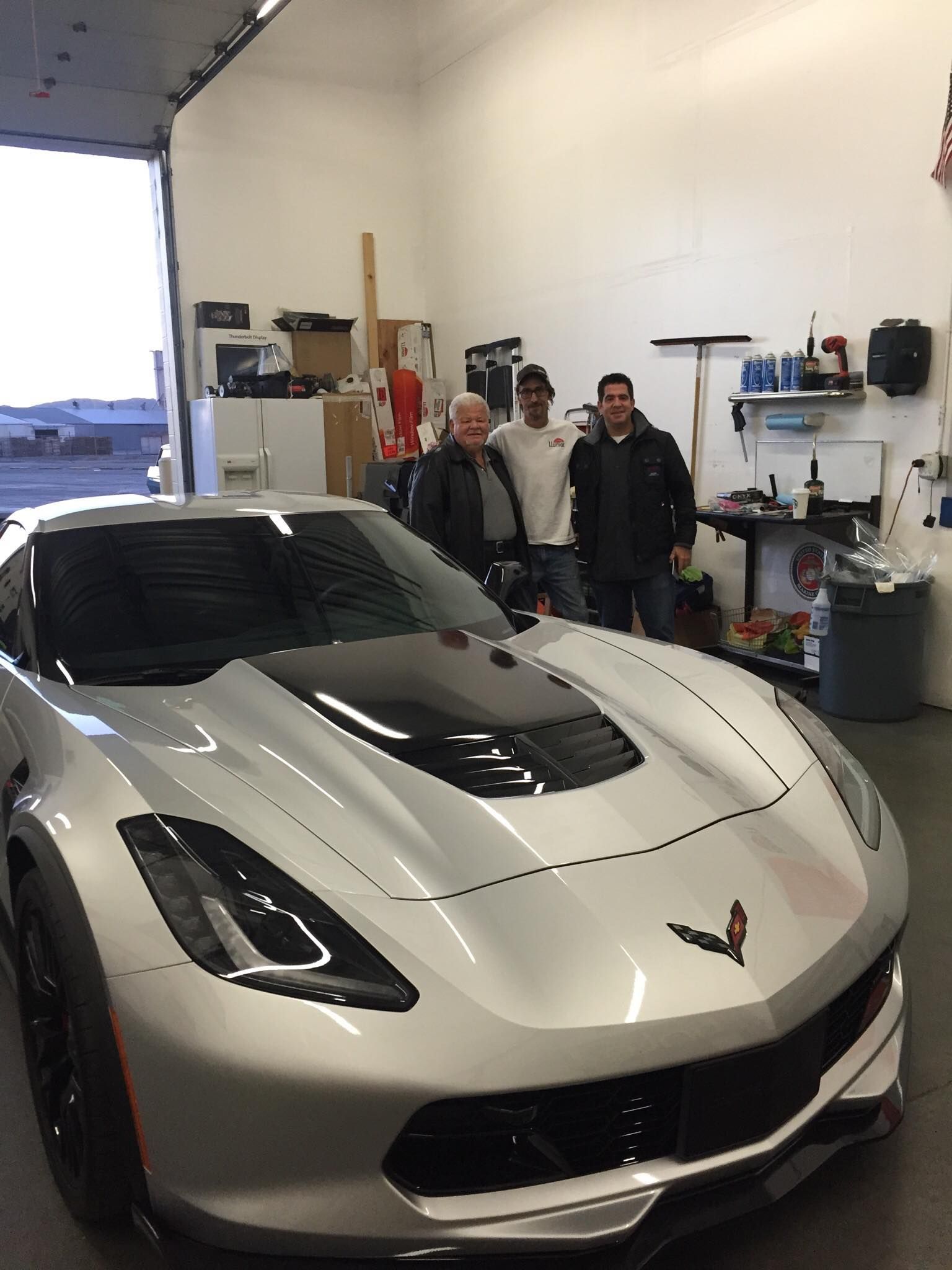Three men are standing next to a silver sports car in a garage.