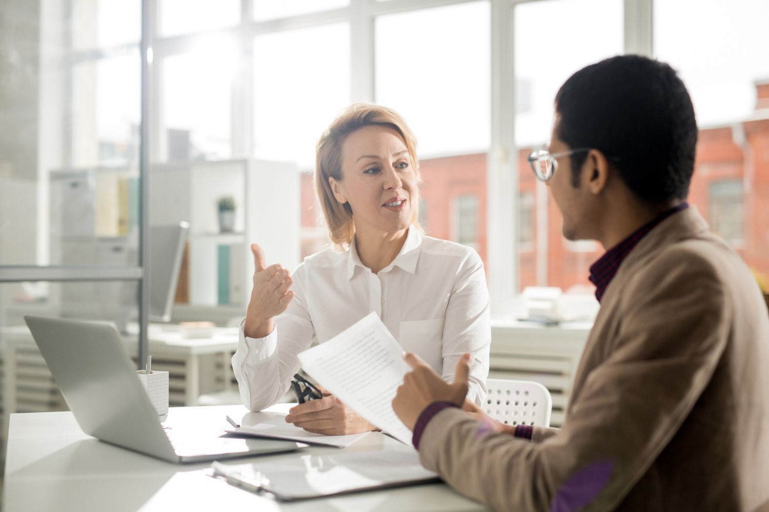 Man in a suit and woman in a shirt sit at a table, looking at each other. There is a laptop and documents on the table.
