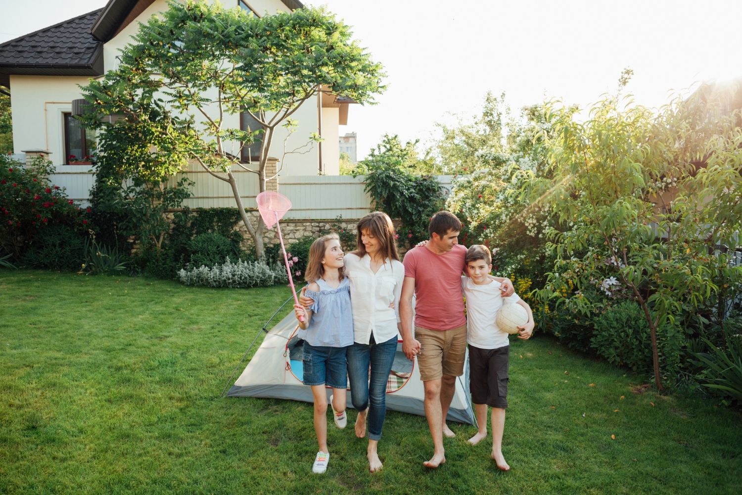 Family walking barefoot on grass toward viewer, tent and house in background.
