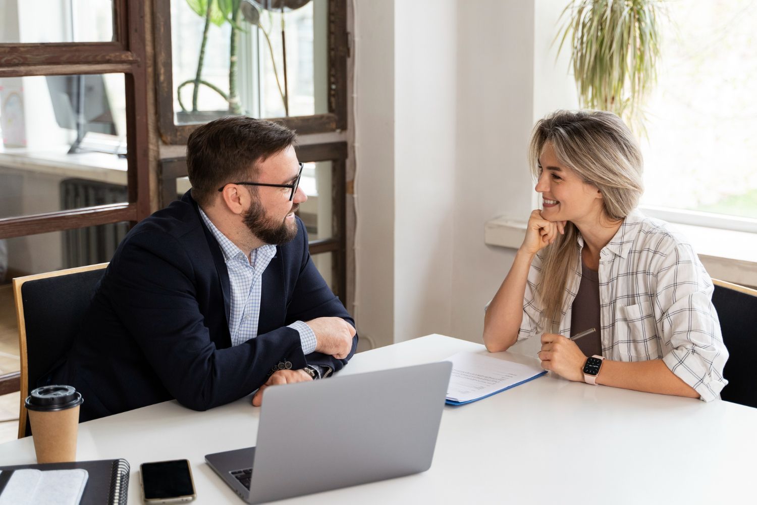 Man in a suit and woman in a shirt sit at a table, looking at each other. There is a laptop and documents on the table.