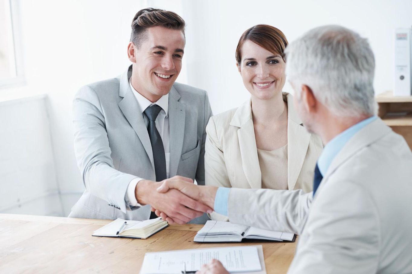 Two people shaking hands at a table, smiling at a third person, in an office.