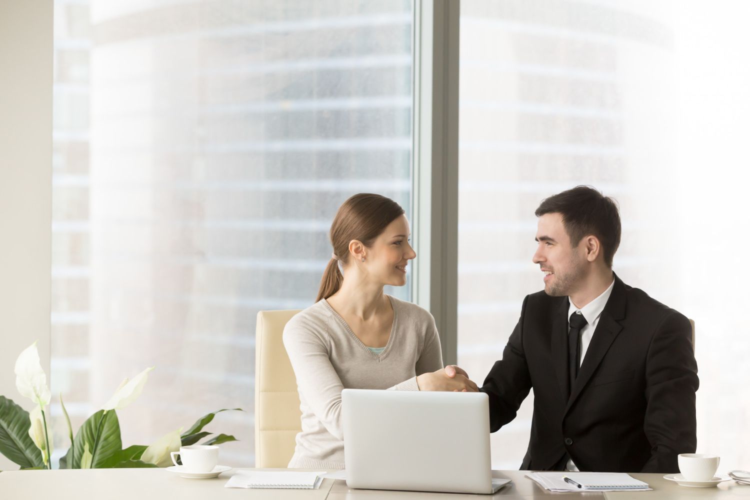 Man in a suit and woman in a shirt sit at a table, looking at each other. There is a laptop and documents on the table.