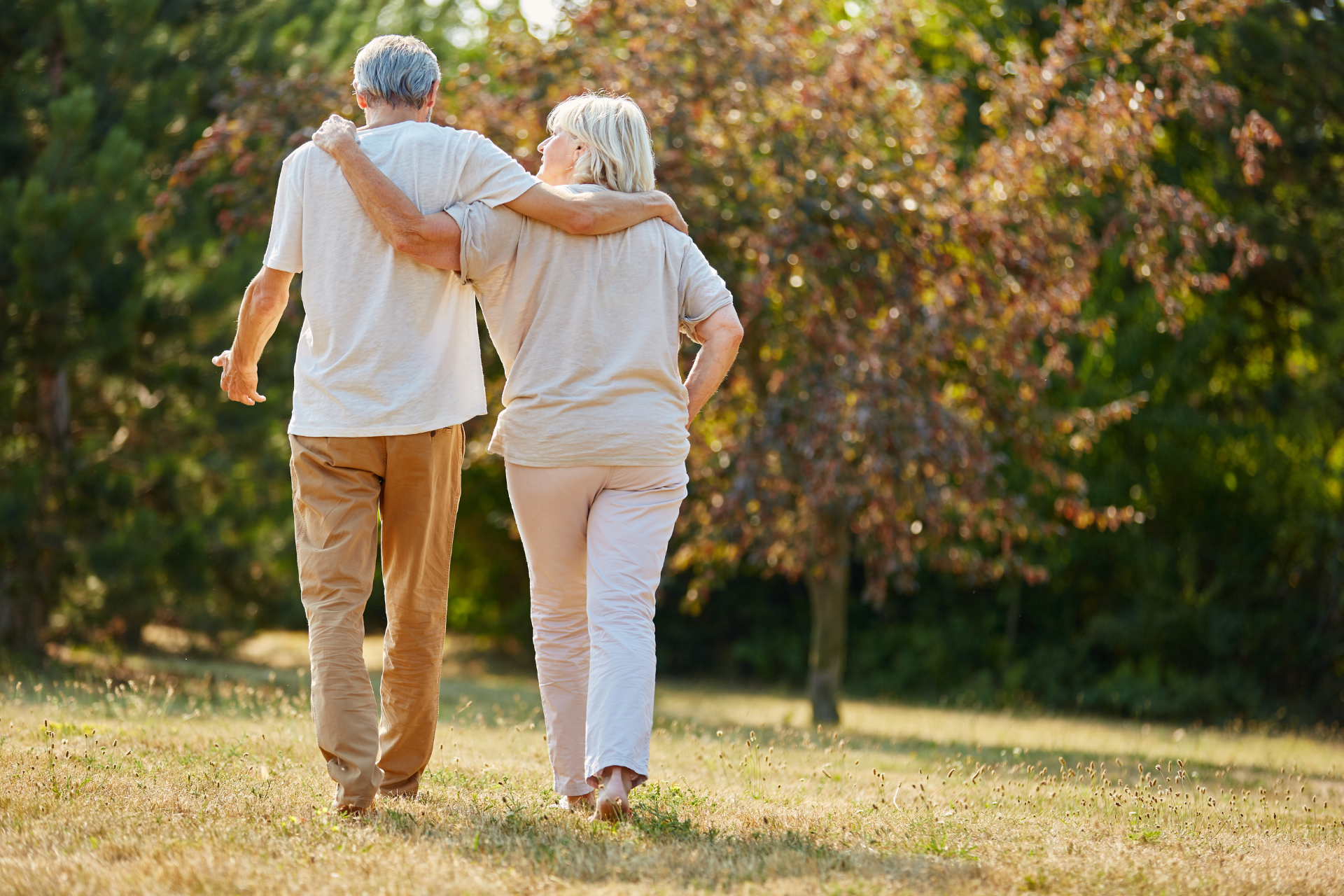 An elderly couple is walking in a park with their arms around each other.