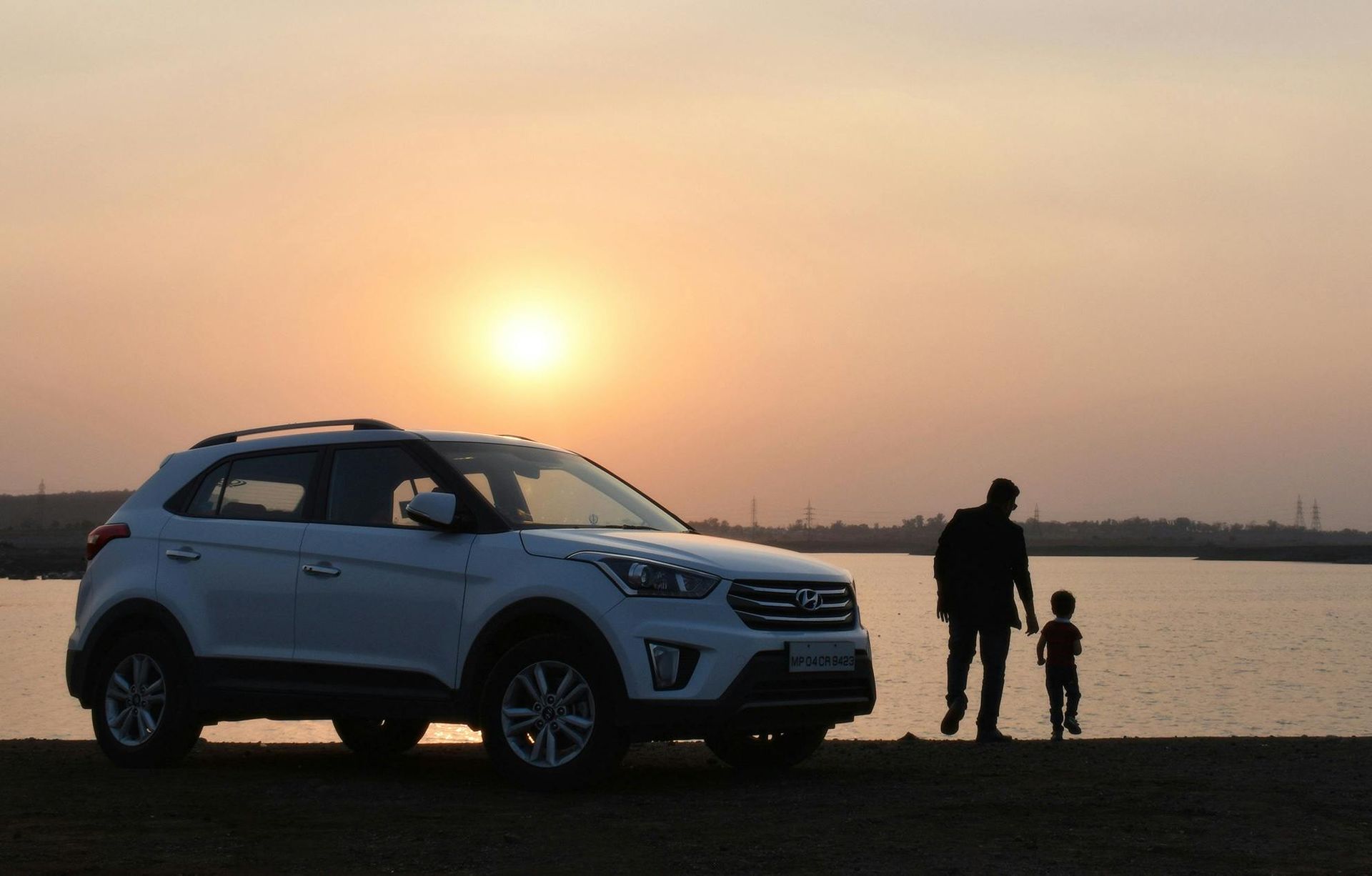 White SUV parked by a lake at sunset; a person and child stand nearby, silhouetted.