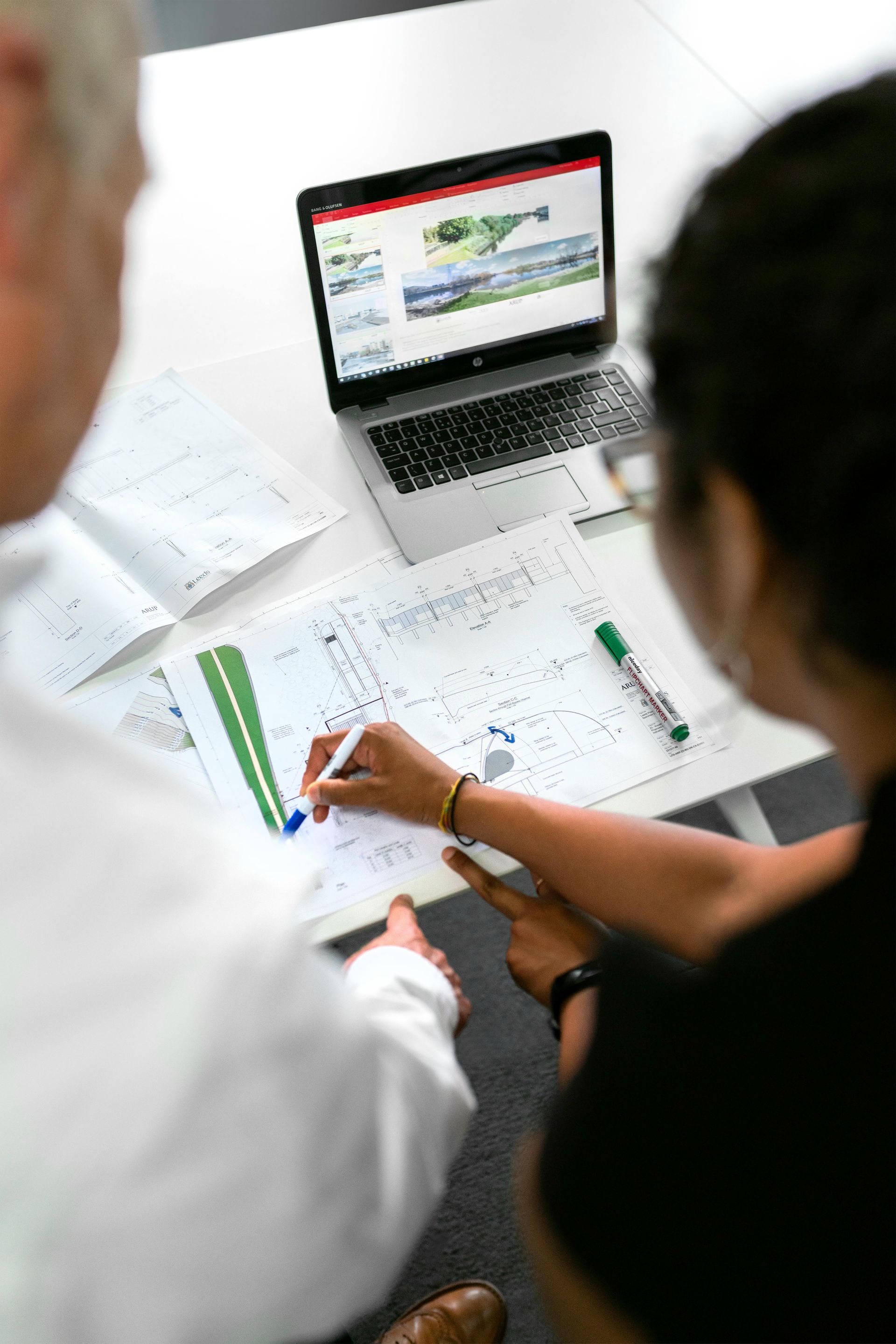 Two people collaborate over technical plans and a laptop on a white desk.