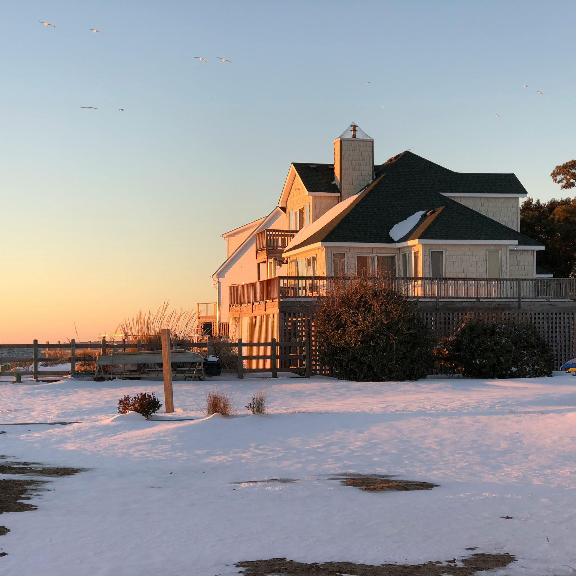 Snowy scene of a house with a green roof, fence, and deck against an orange and blue sky.