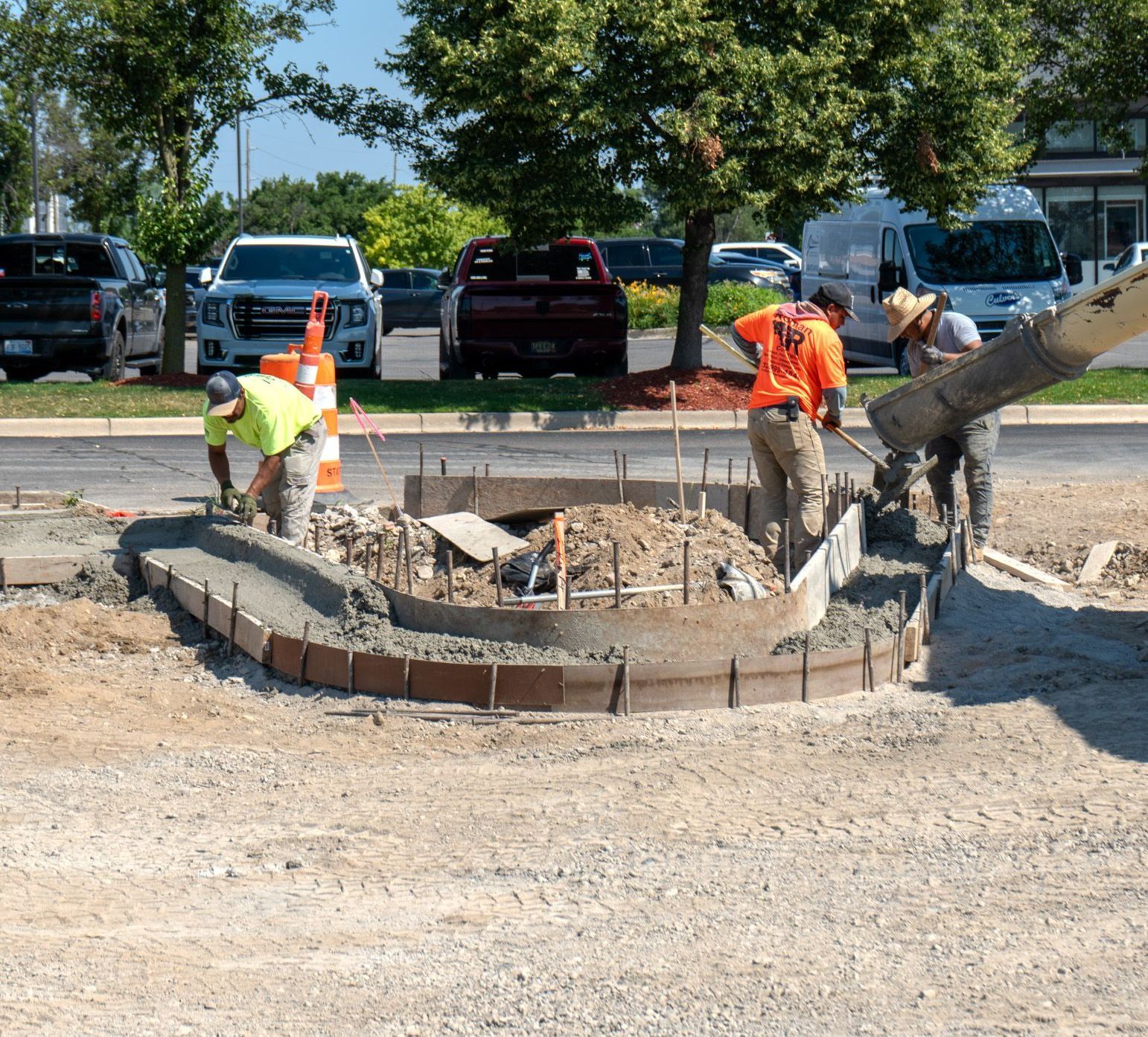 Construction workers pouring concrete for a circular curb in a parking lot, orange safety vests, sunny day.