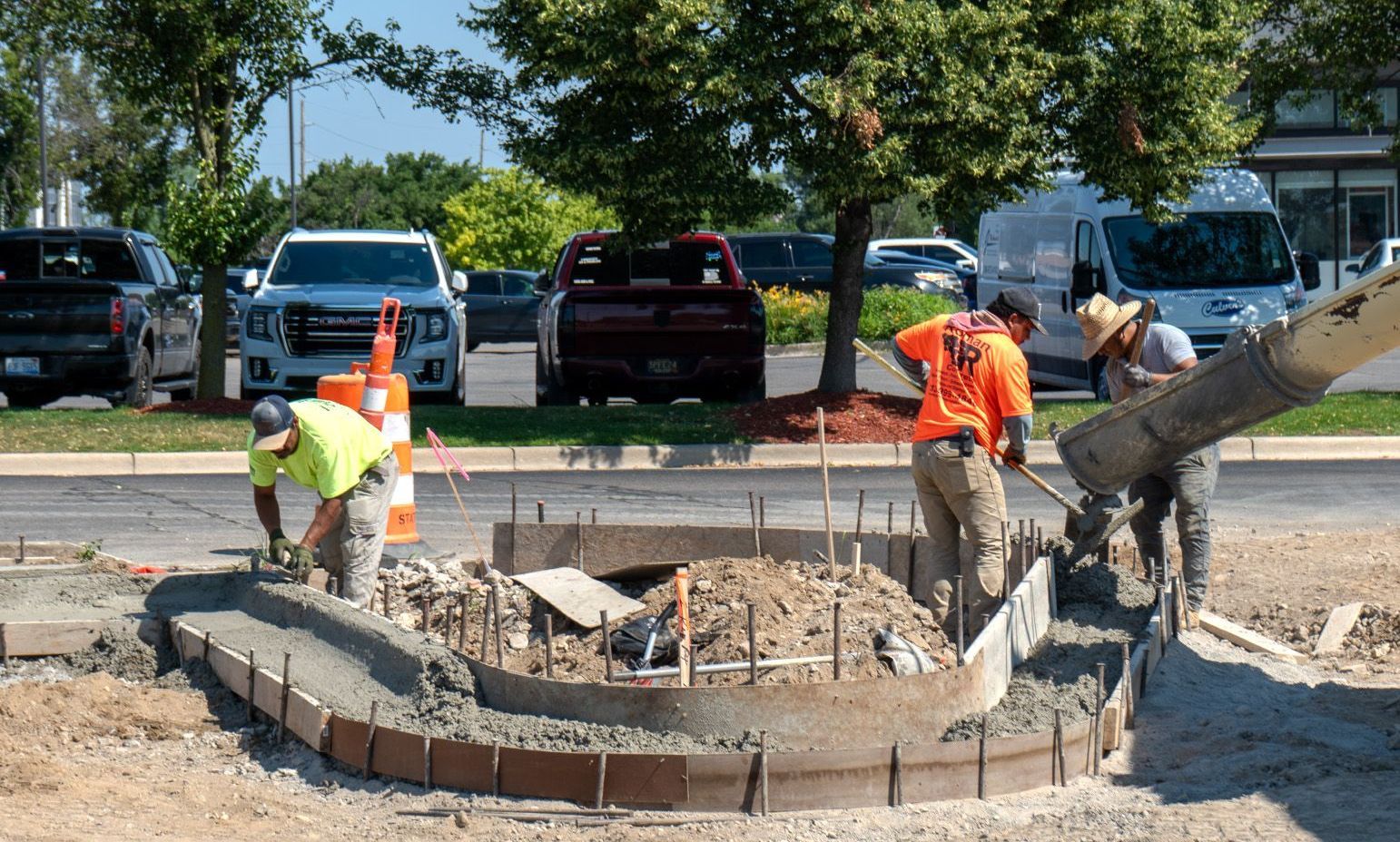 Construction workers pouring concrete for a curved flower bed; vehicles and trees in the background.