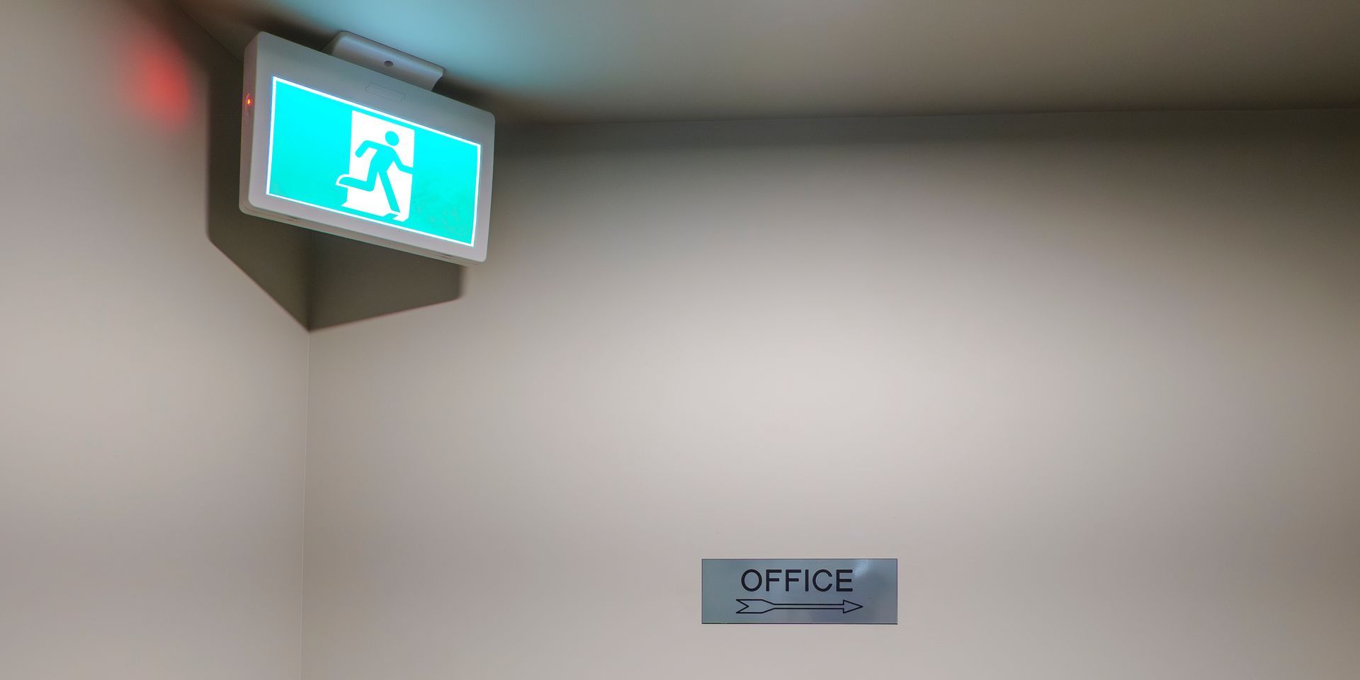 Illuminated exit sign hangs above a hallway wall with an office directional sign below.