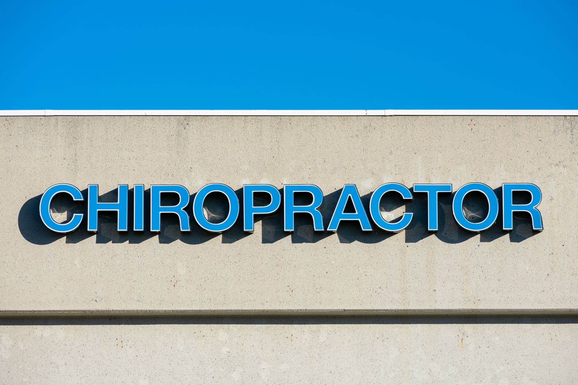 Chiropractor clinic sign on beige wall, bold blue letters under clear sky.