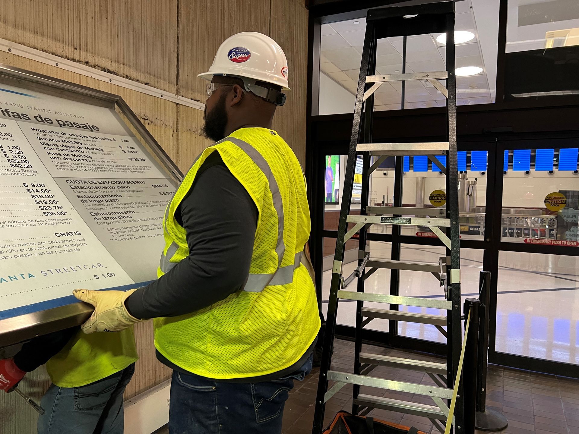 A man in a yellow vest and hard hat is standing next to a ladder holding a sign.