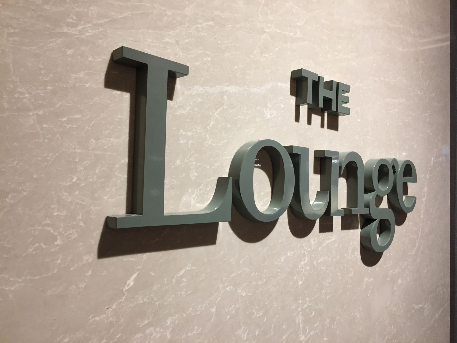 3D sign reading “THE Lounge” mounted on light-textured wall, casting shadows below letters.