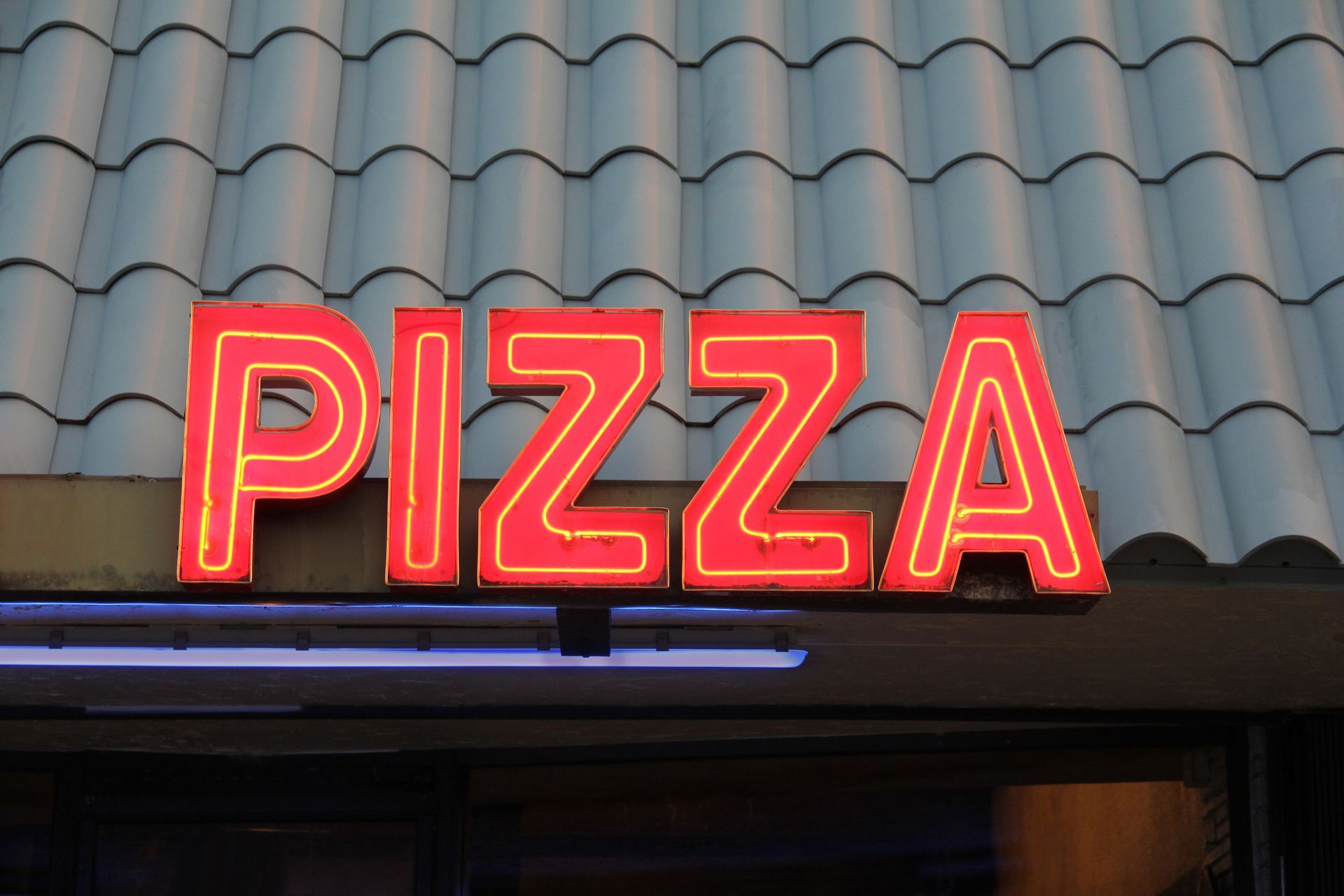 A bold red neon 'PIZZA' sign on a blue-tiled roof—an example of commercial exterior signs.
