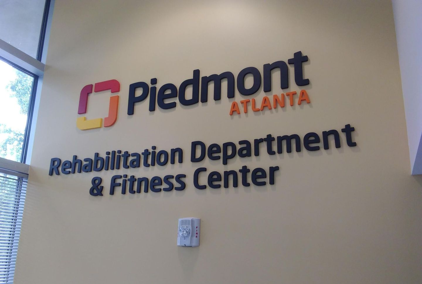 Sign for Piedmont Atlanta Rehabilitation Department & Fitness Center. Red and orange logo, black text on beige wall.