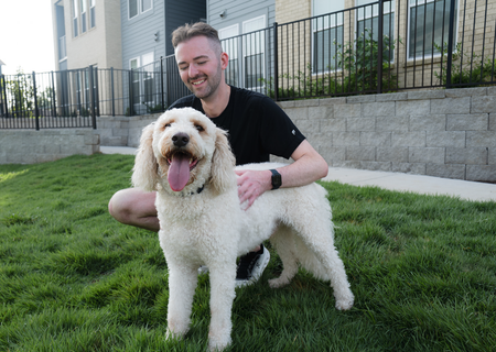 Man kneels on grass beside a fluffy white dog in front of an apartment building