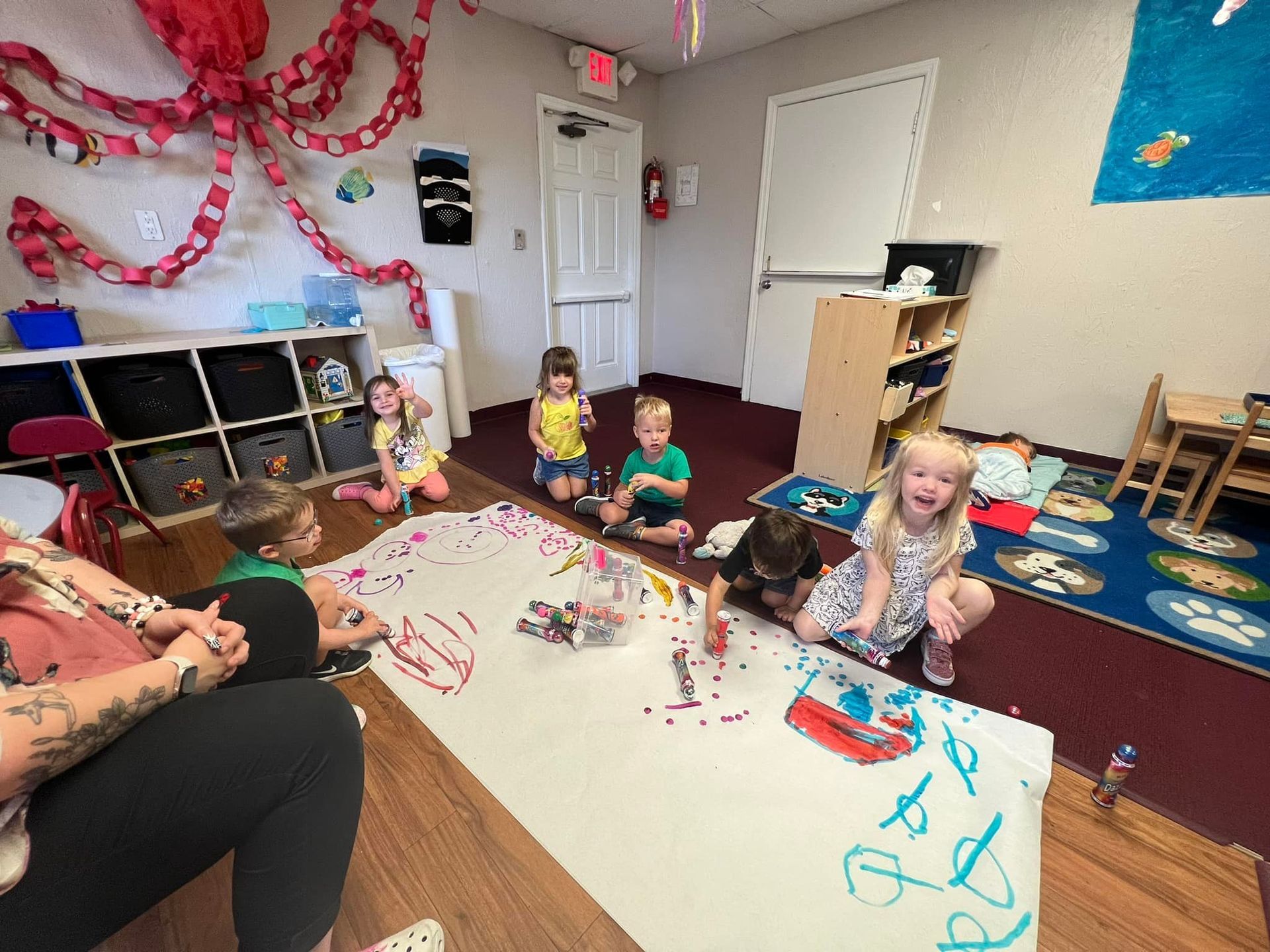 Children painting on a large white sheet in a classroom, supervised by an adult. Octopus decoration overhead.