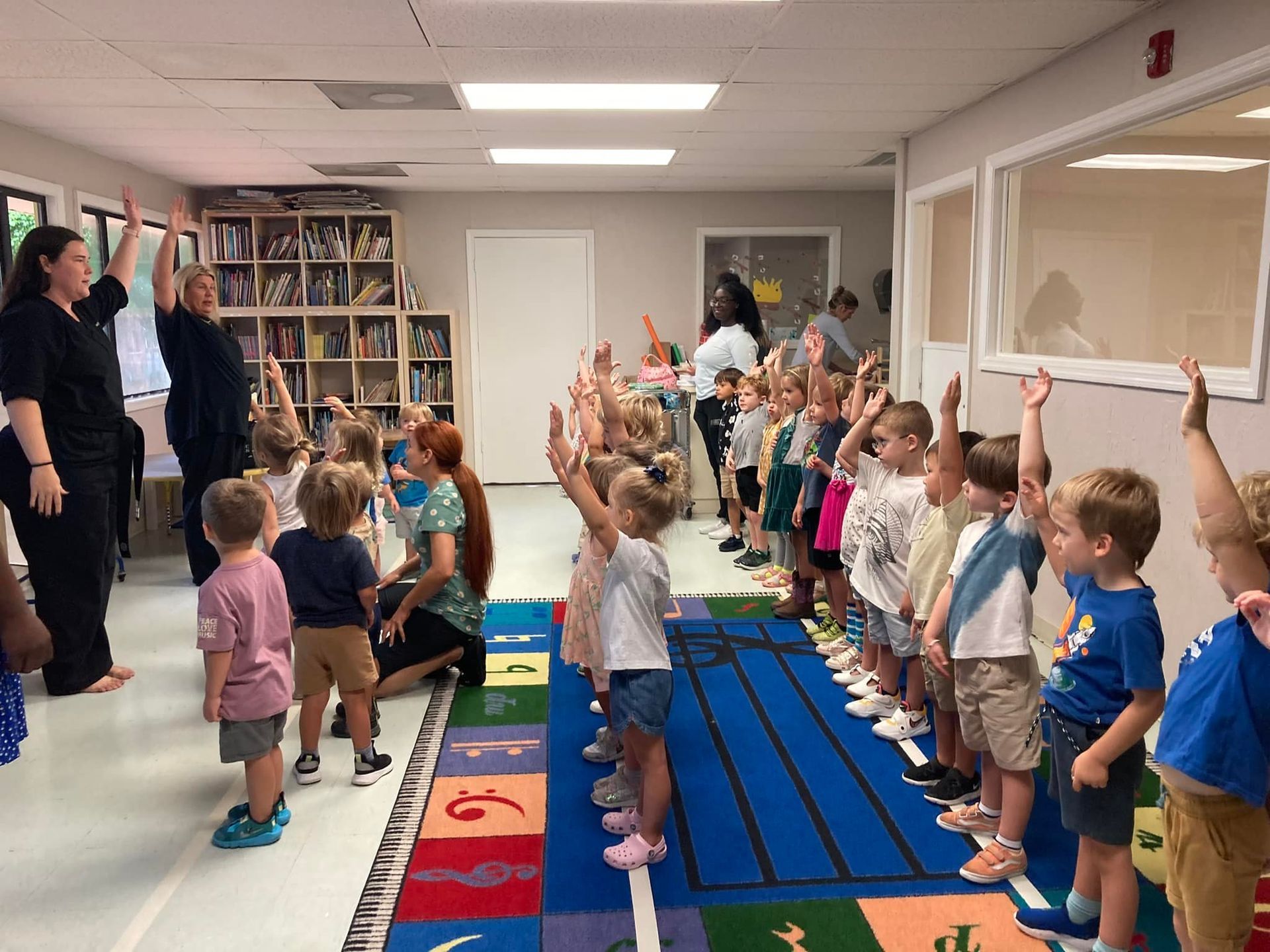 Children and adults in a classroom raise arms. Bookshelves and rug with music notes are visible.