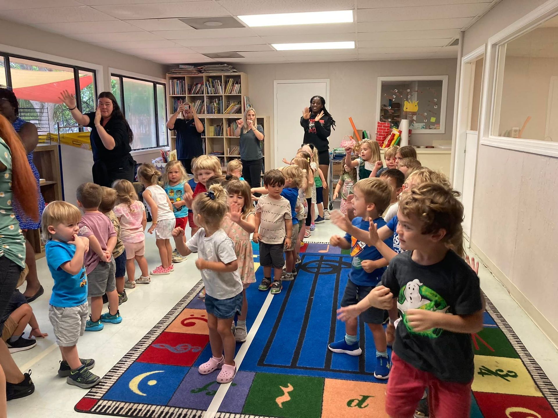Children and adults clapping in a classroom. Blue carpet, bookshelves, and windows are visible.