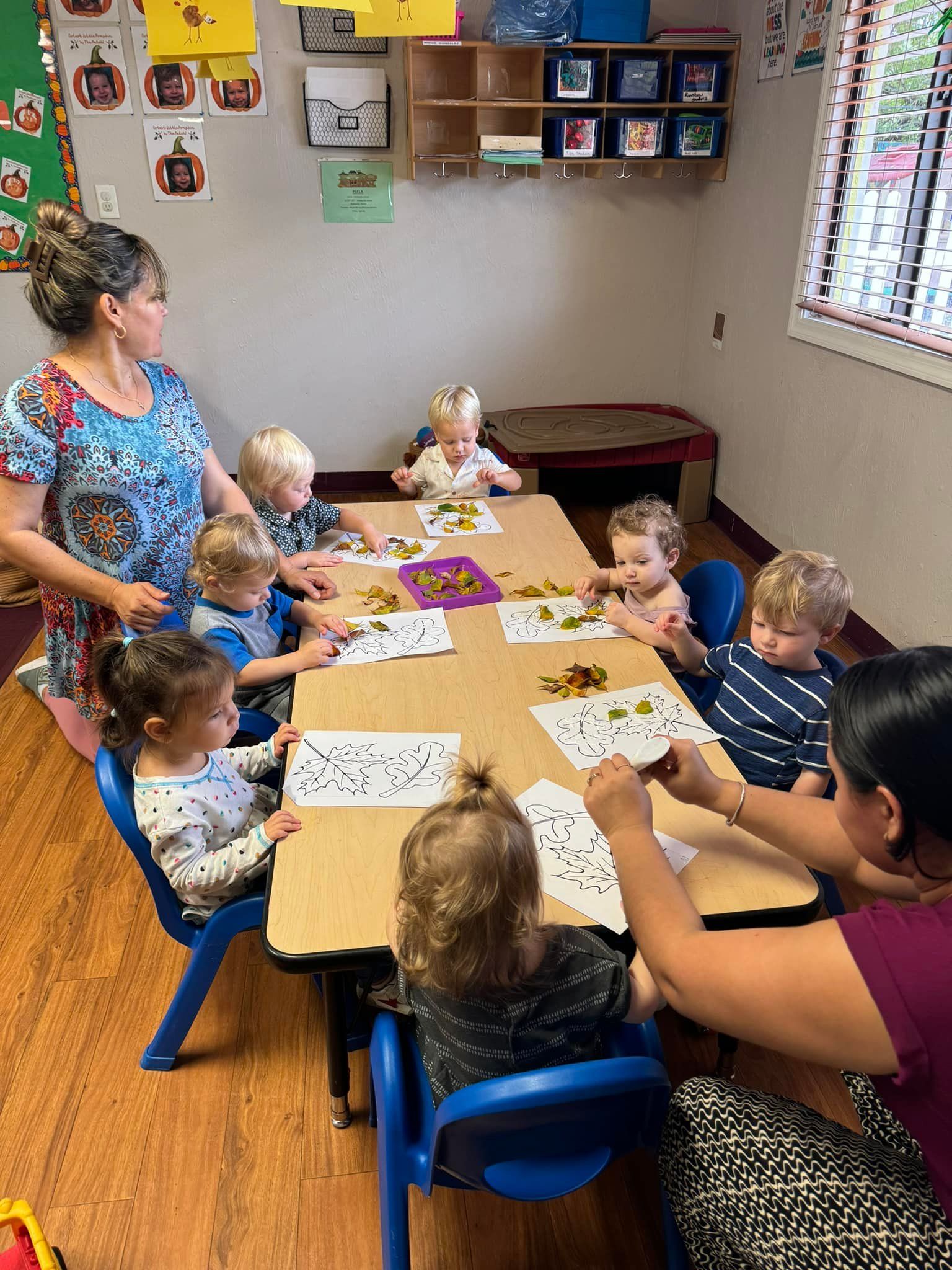 Children and adults at a table, crafting with paper and greenery in a classroom setting.