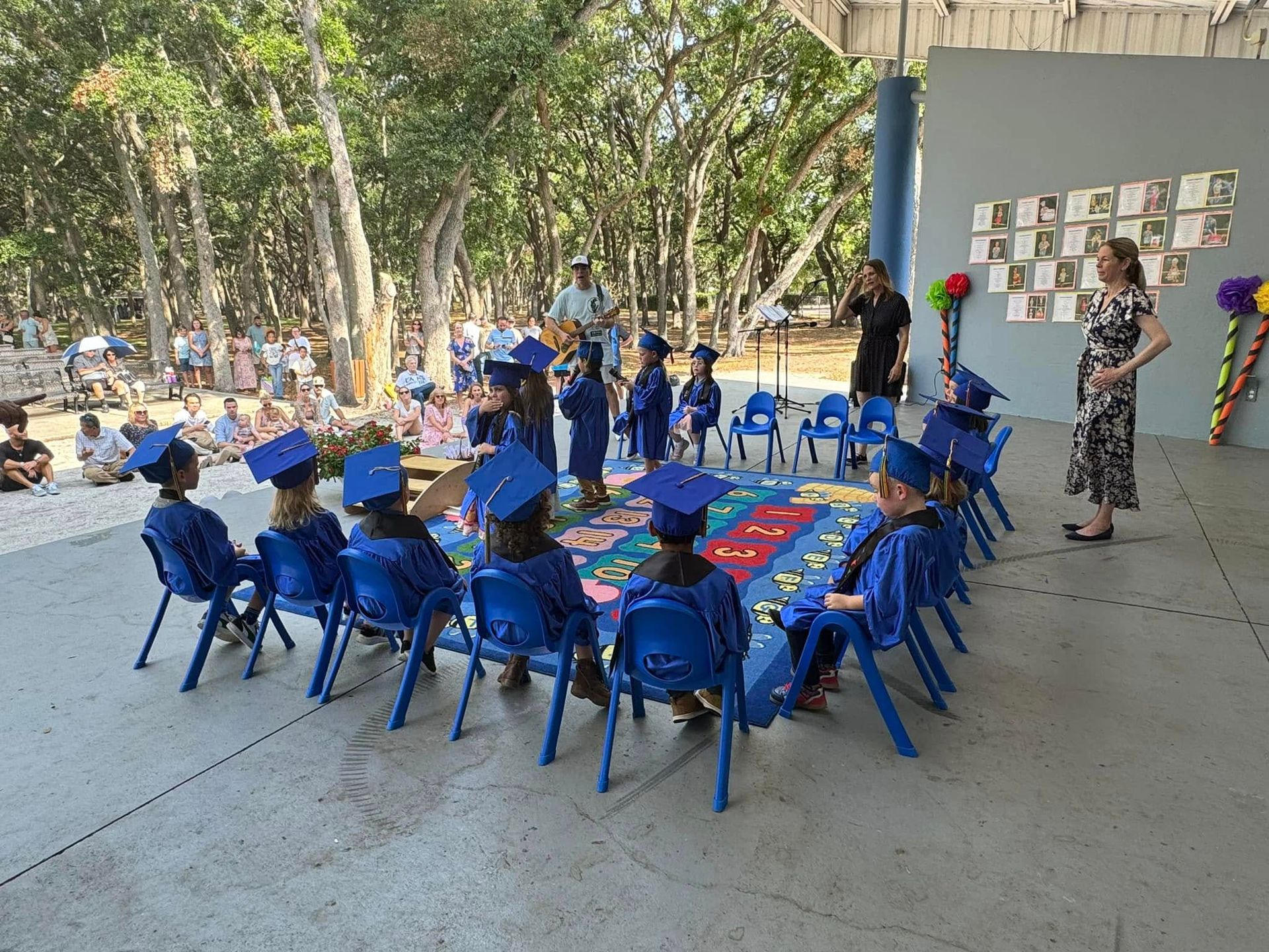 Children in blue graduation gowns and hats at an outdoor ceremony, with adults and a decorated backdrop.