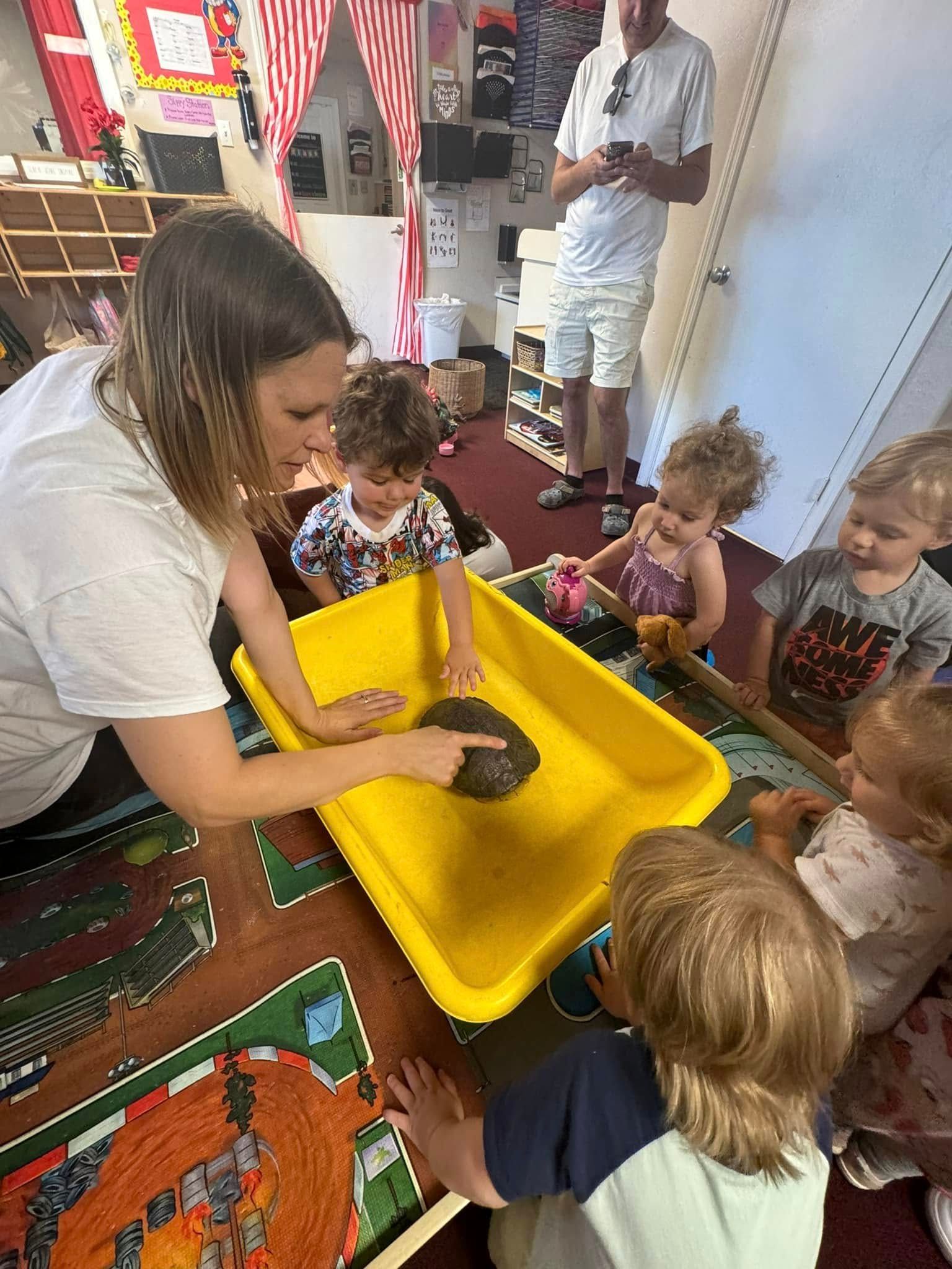 A teacher points to something in a yellow bin. Children observe. White shirt, classroom setting.