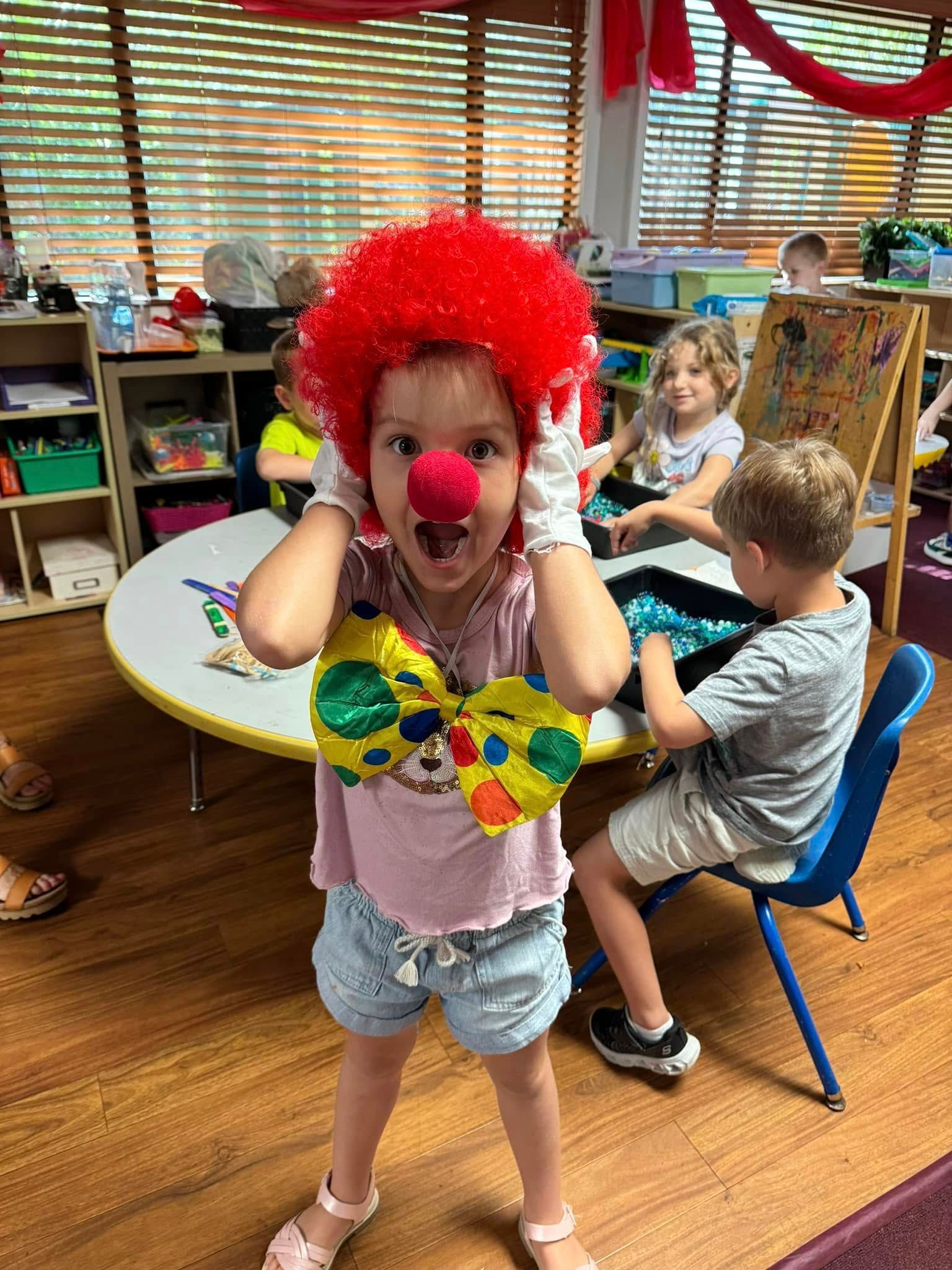 Child in clown costume with red wig, nose, and bow tie in a classroom.