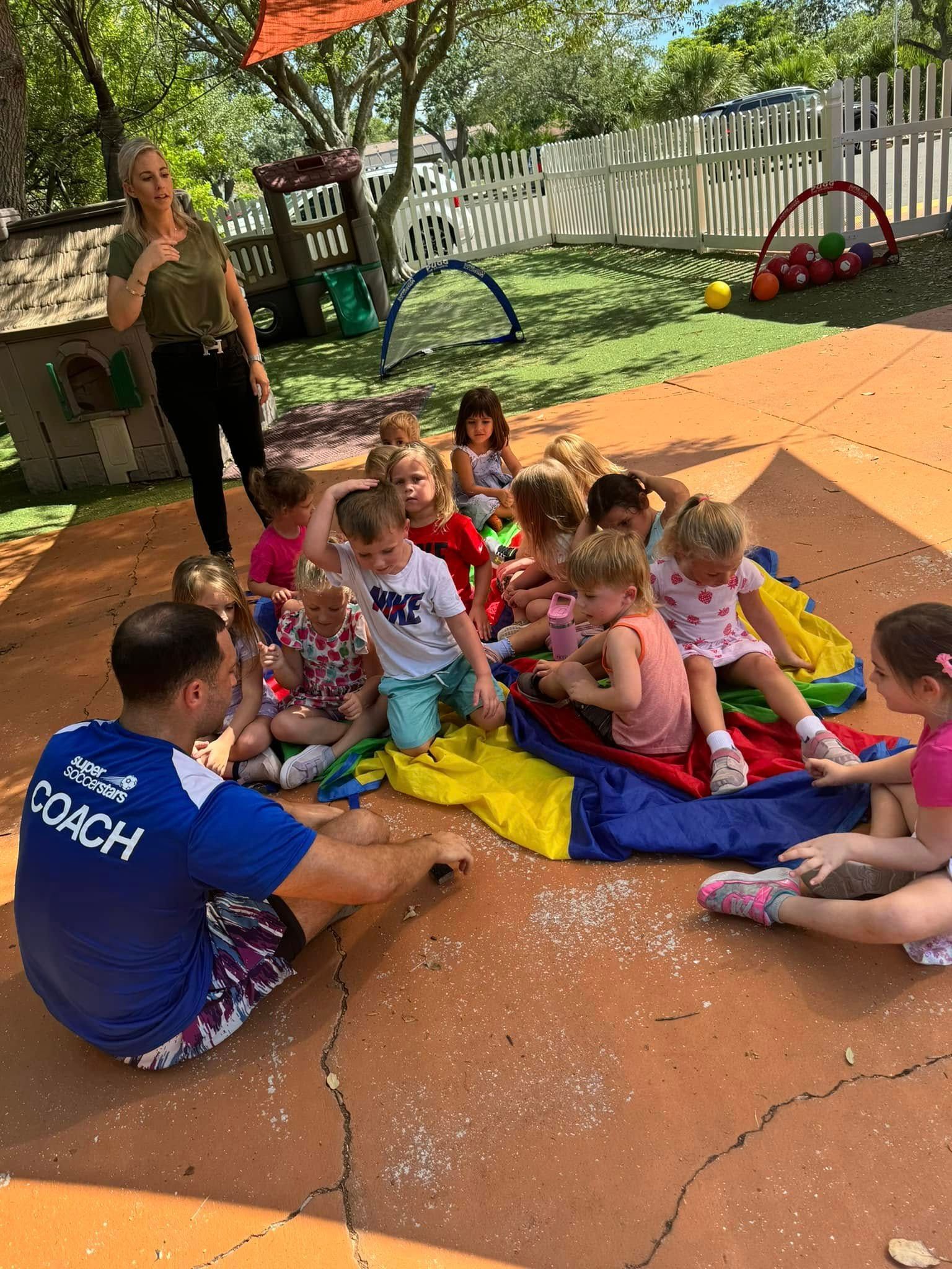 Children and a coach sit on a colorful parachute on a playground. Another adult stands nearby.