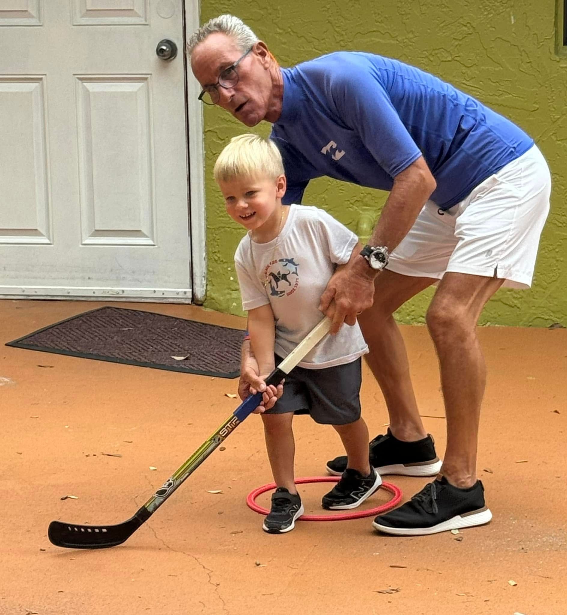 Man assisting a child with a hockey stick and a red hoop on a patio.