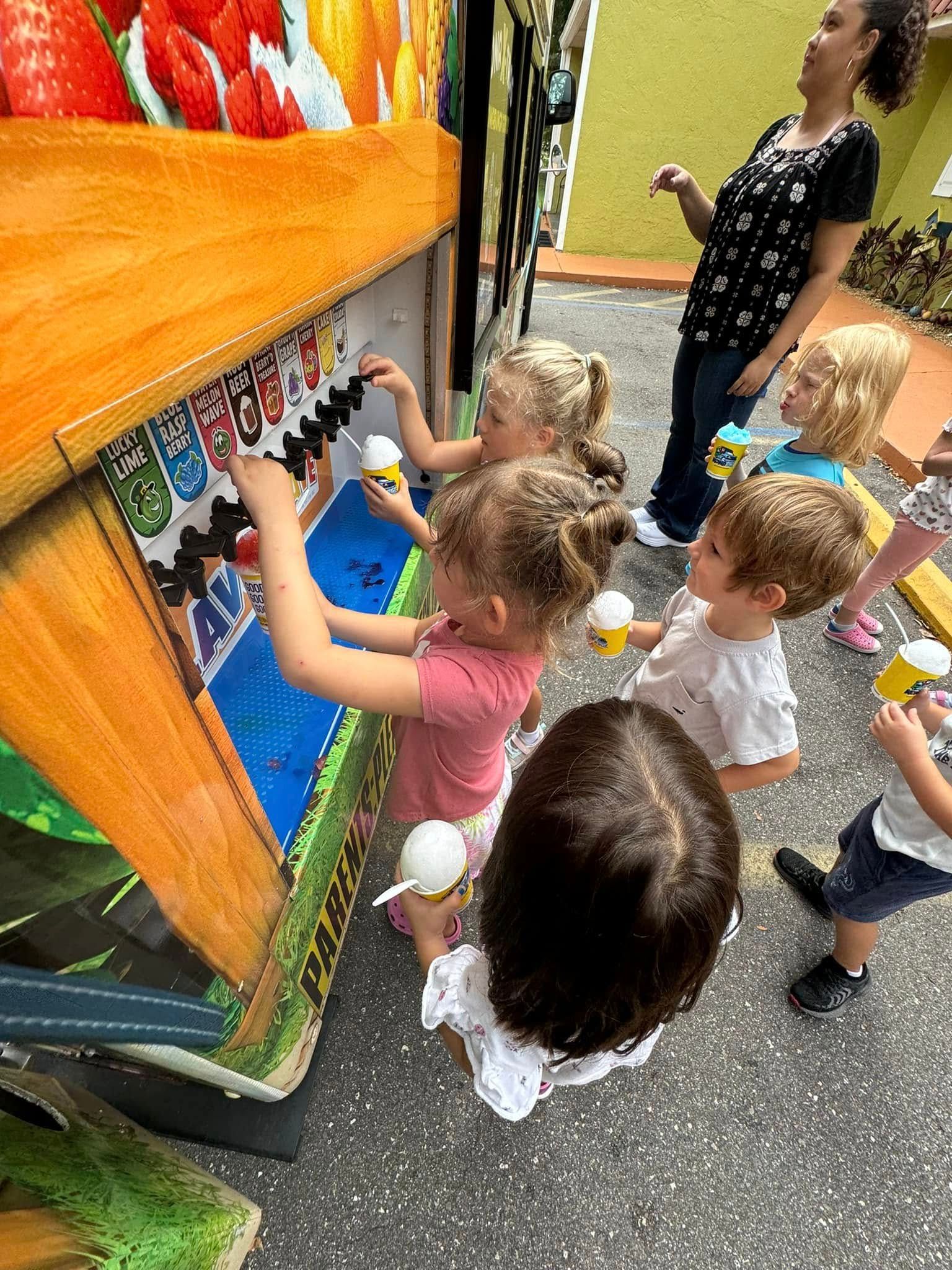 Children interacting with a colorful toy, with a woman observing. Outdoors, sunny day.