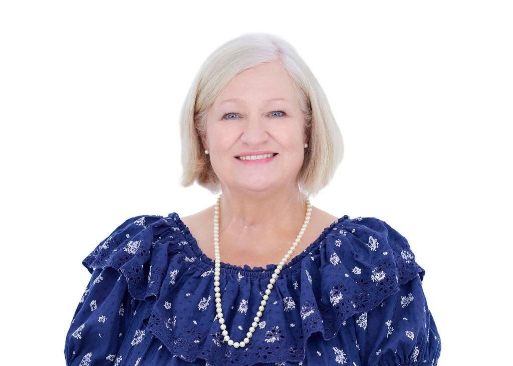 An elderly woman wearing a blue top and a pearl necklace is smiling.