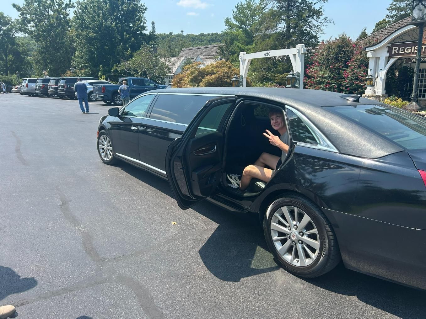 Black limousine with an open door. A person waves from inside. Outdoors, sunny.