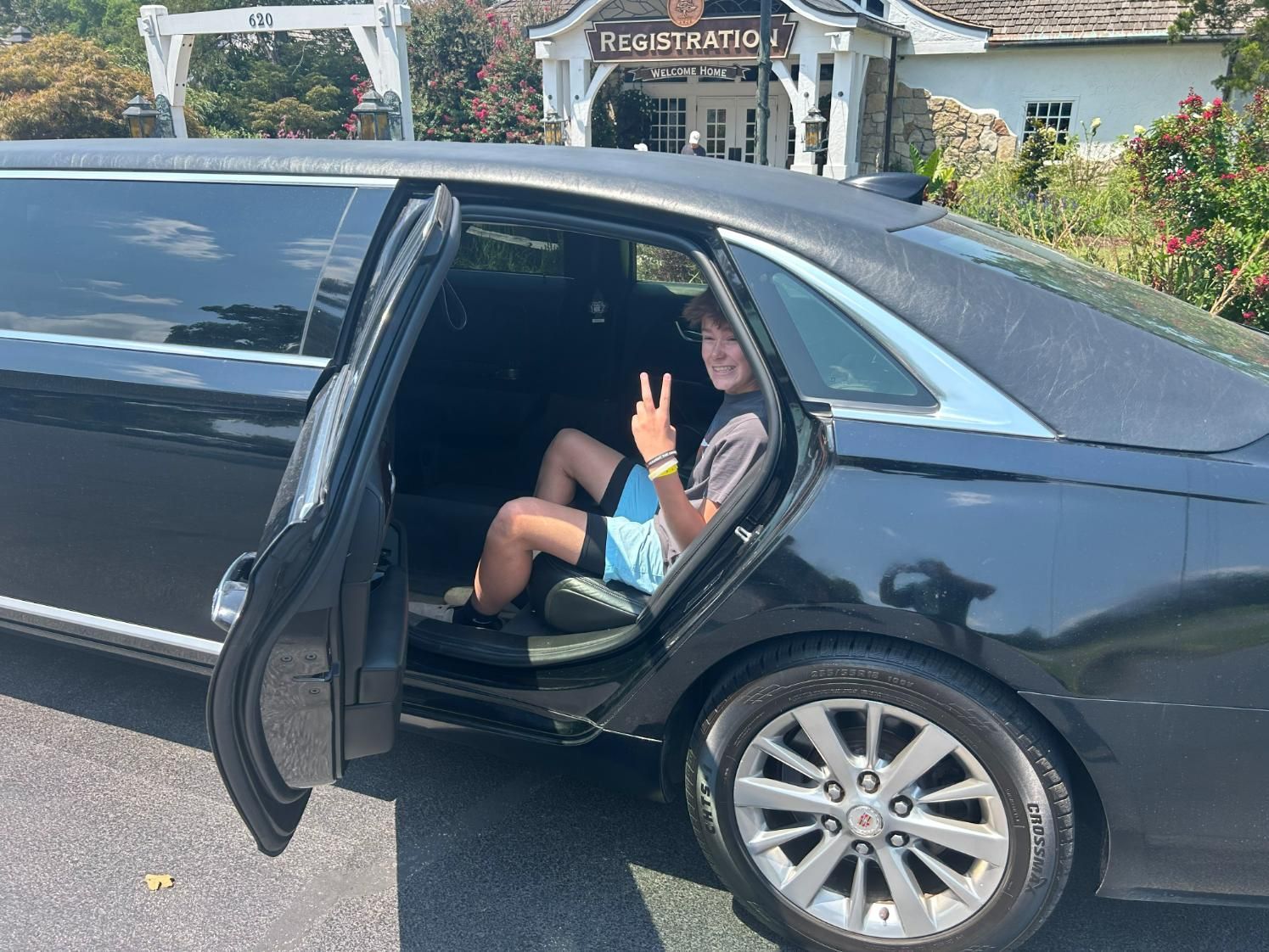Boy in shorts giving a peace sign, sitting in a black limousine with door open in front of a building.