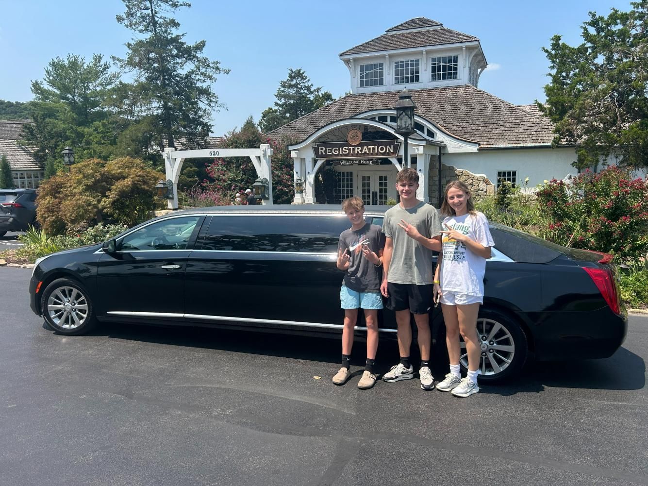 Three young people pose next to a black limousine in front of a building with a sign, 