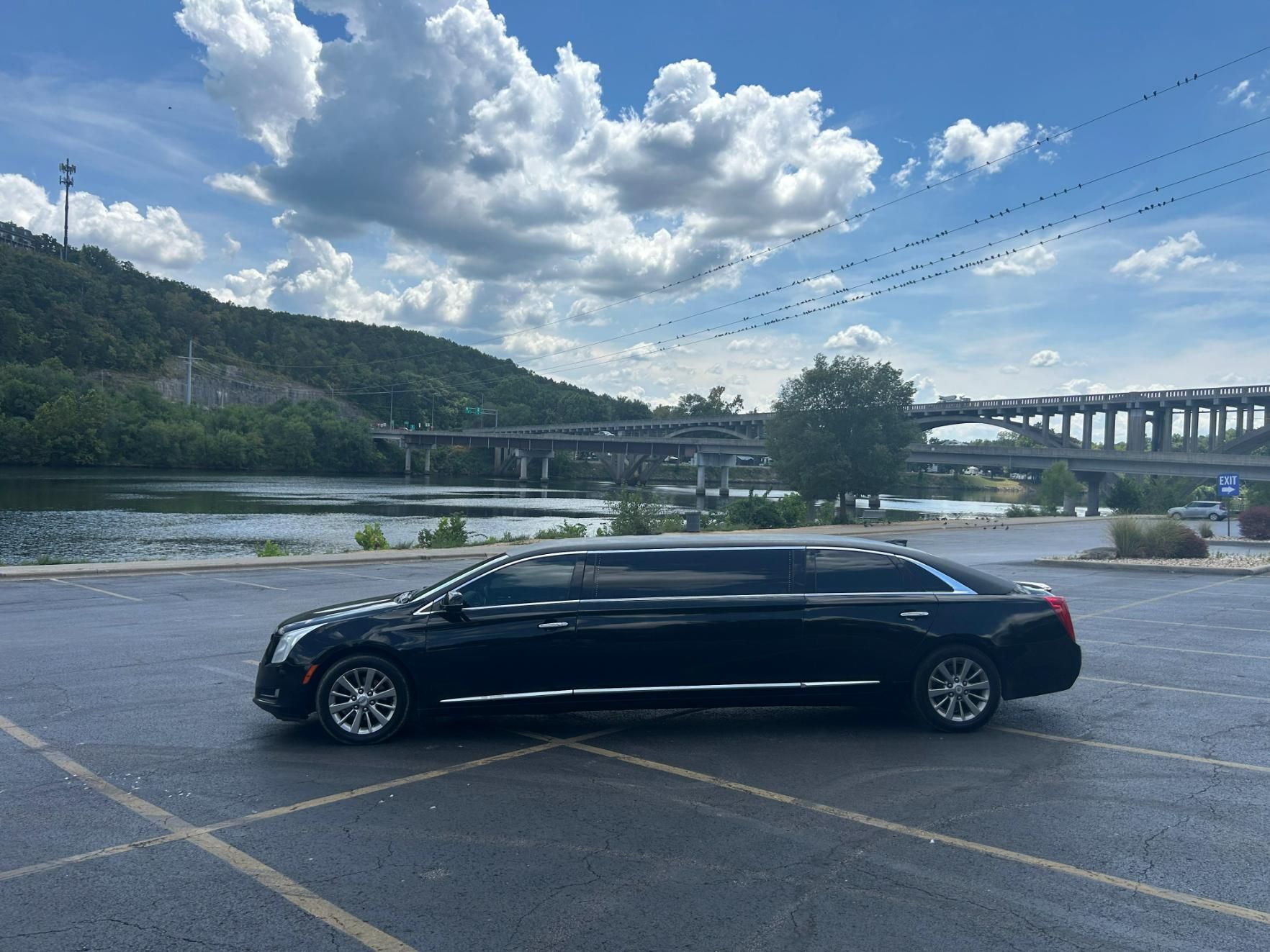 Black limousine parked by a river with a bridge in the background under a cloudy sky.