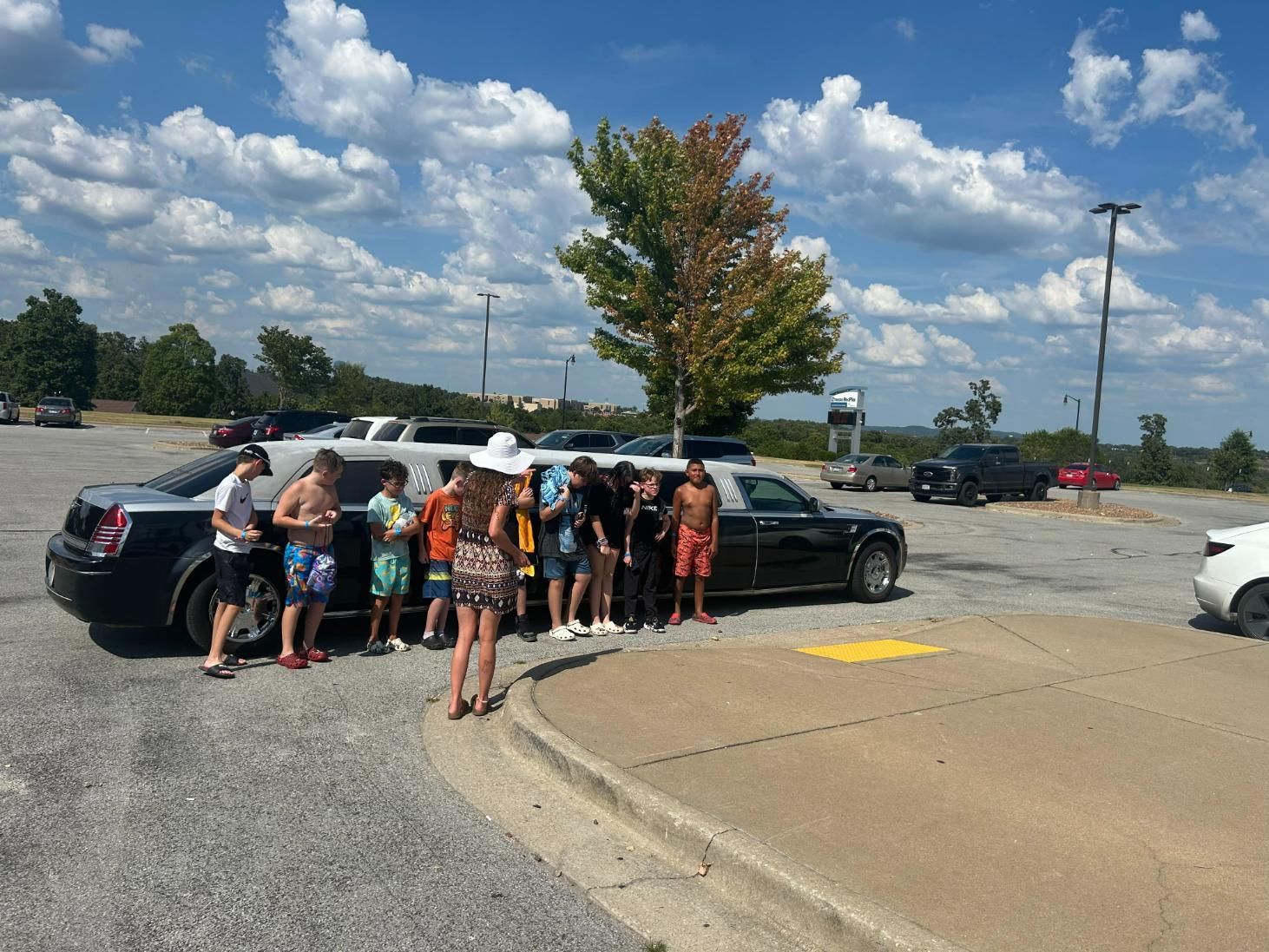 A group of people in swimsuits standing next to a black limousine in a parking lot on a sunny day.