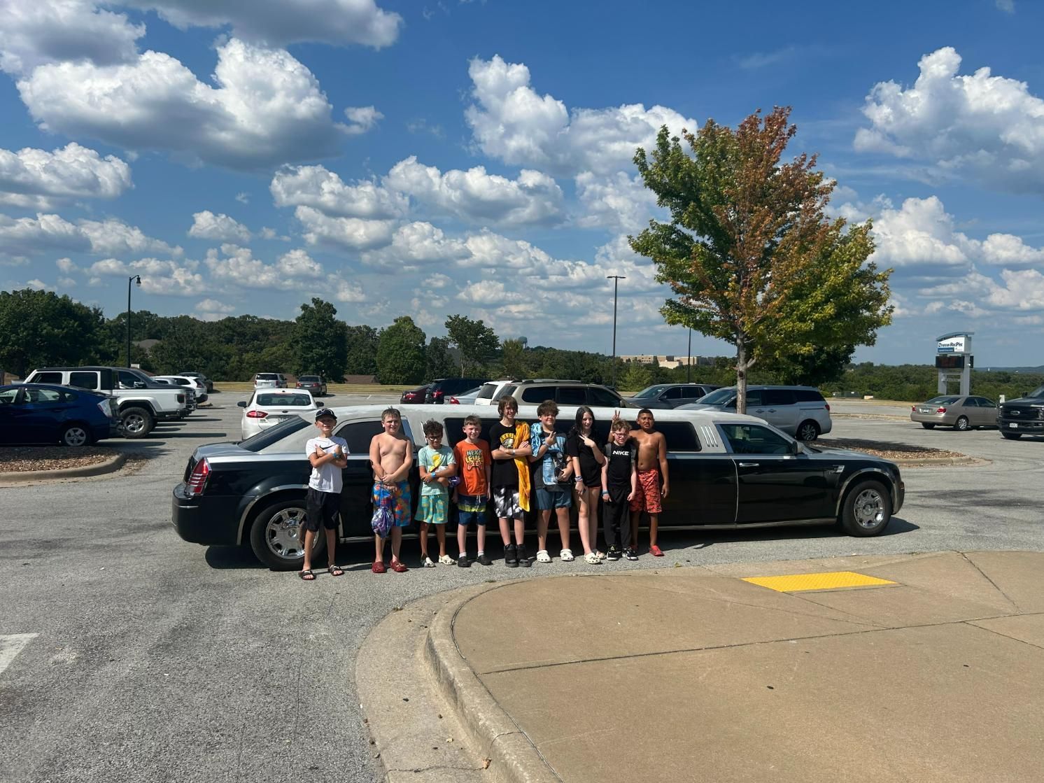 Group of teens poses with a black limousine in a parking lot on a sunny day.